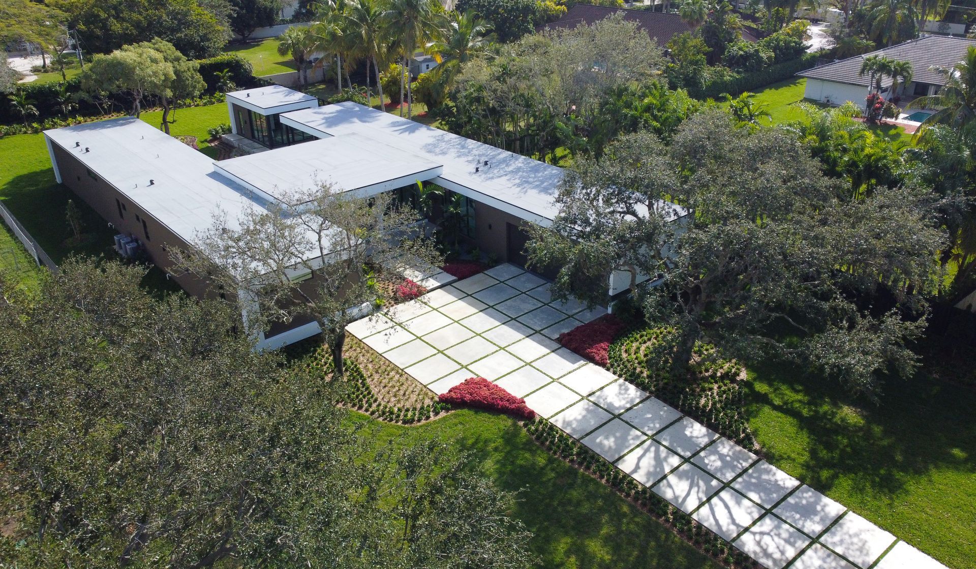 Modern, single-story house with a flat roof. A walkway leads to the entrance. Lush green lawn and trees surround the home.