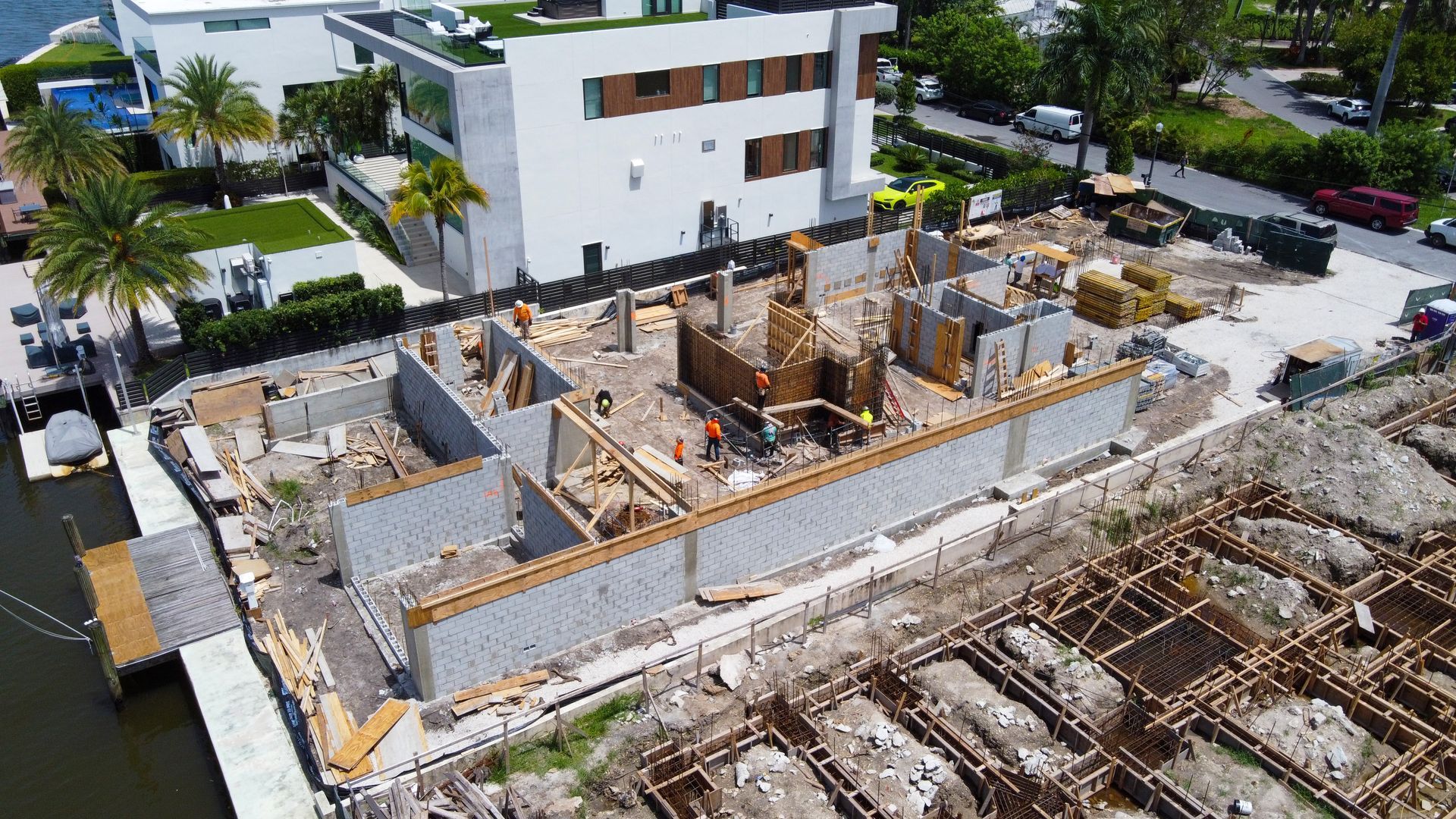 Construction site of a house by the water, with concrete walls, wooden framing, and workers.