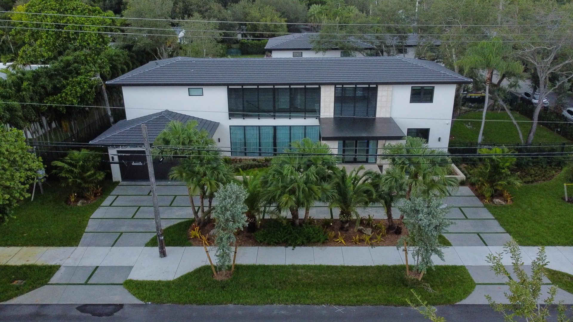 Modern two-story house with black roof and windows, surrounded by trees and a paved driveway.
