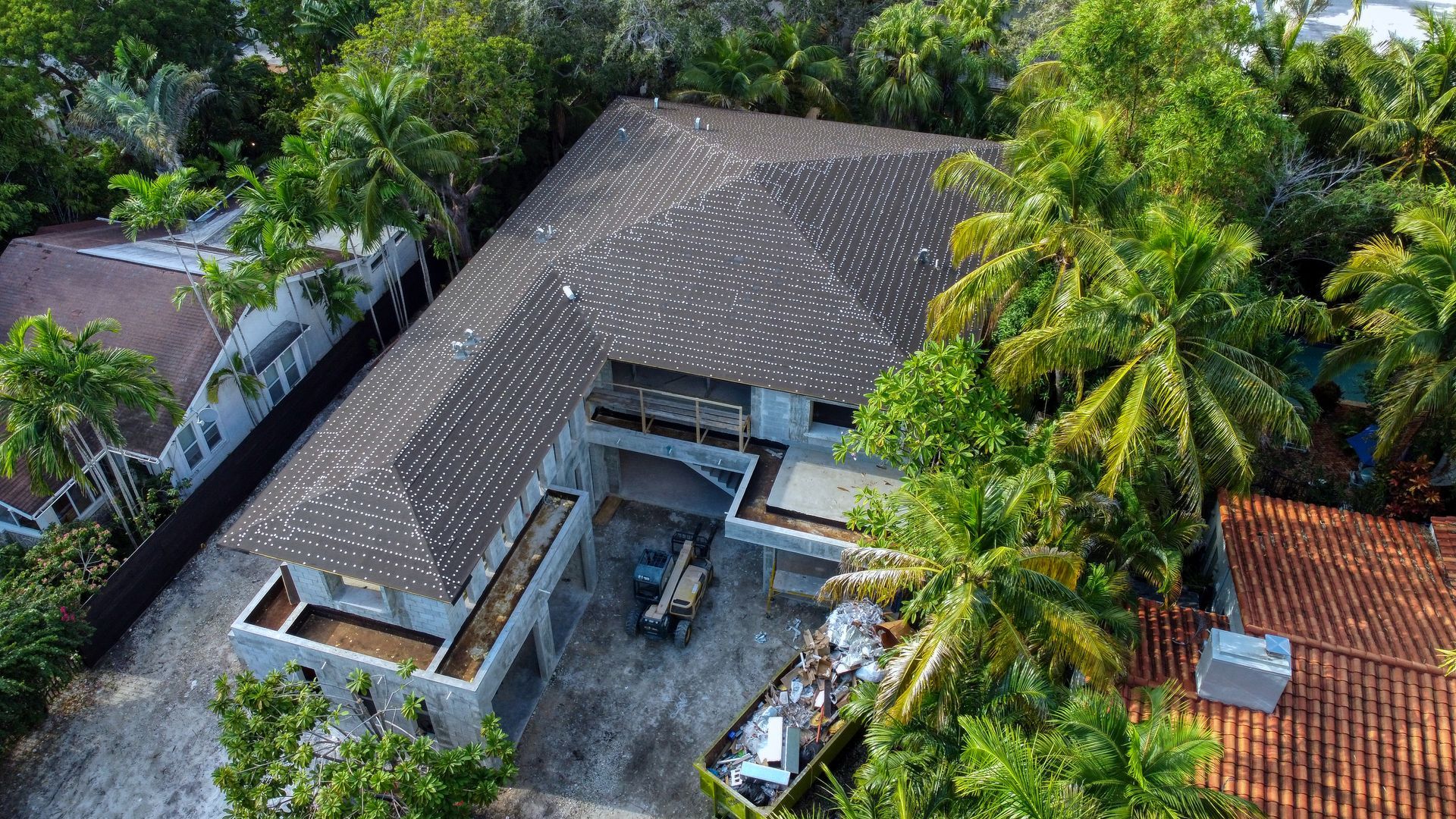 Aerial view of a gray house surrounded by green trees. Construction equipment visible in front.