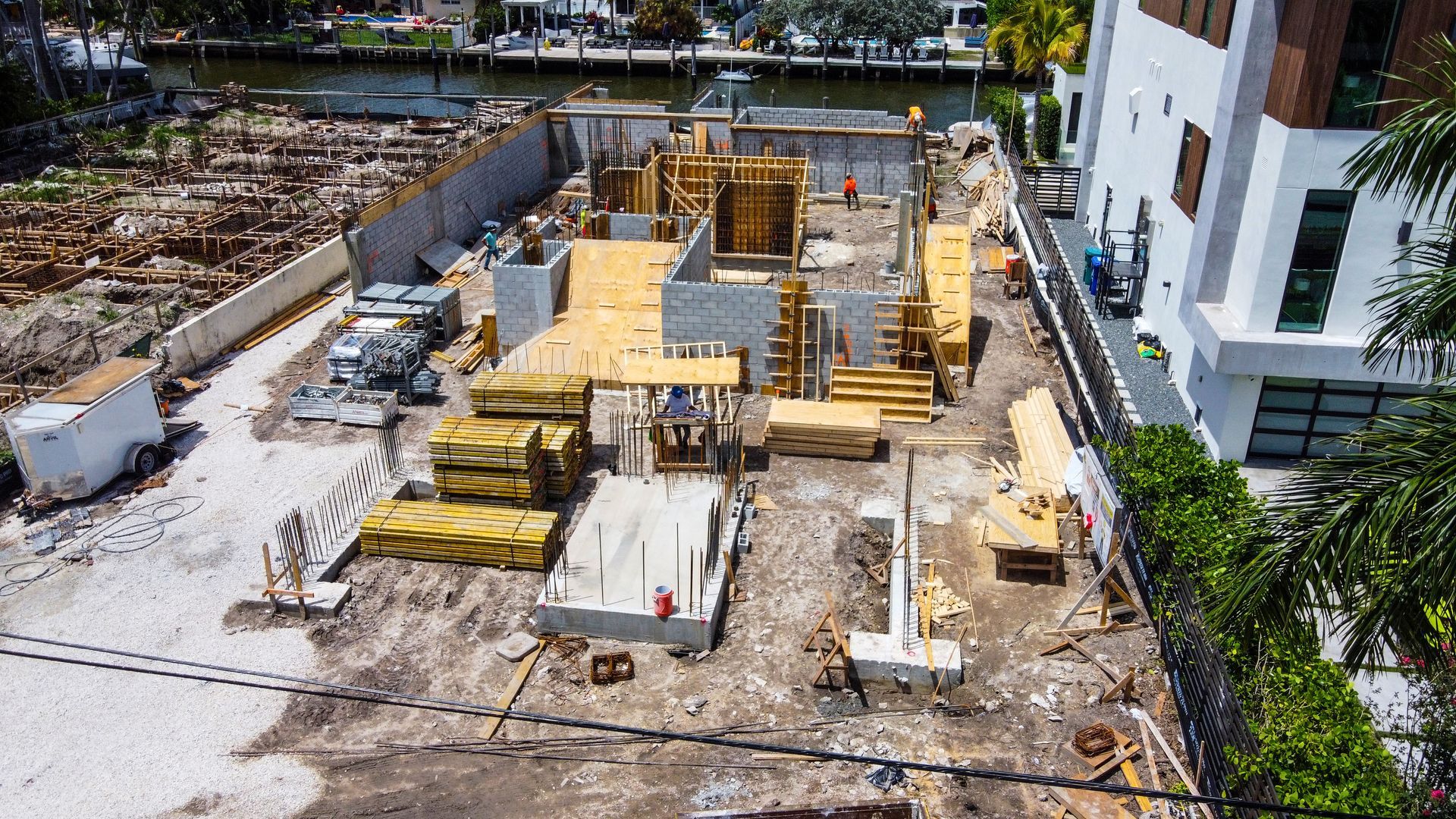 Construction site: foundation and framing in progress, with wood, concrete, and materials, near a waterfront.