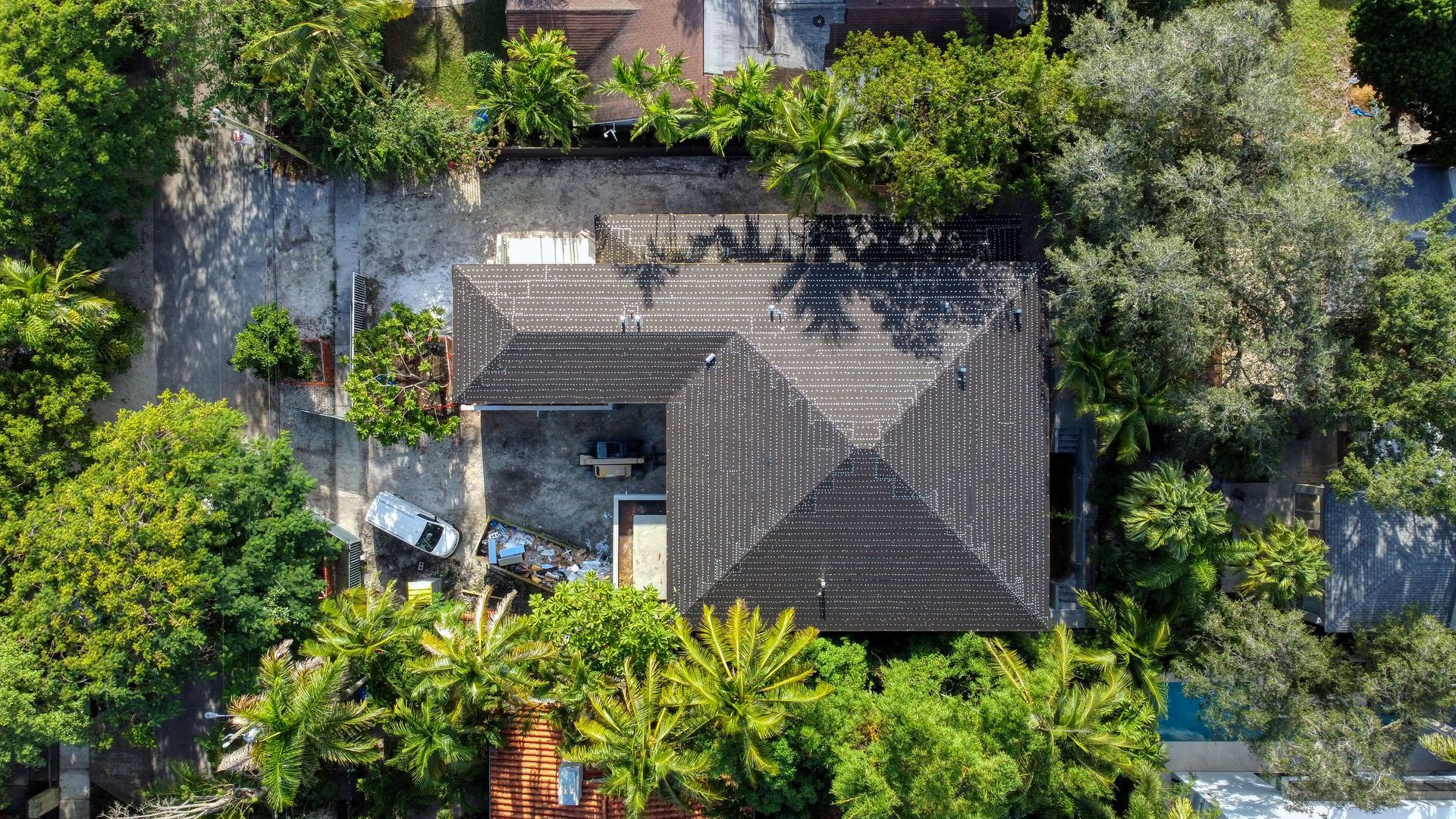 Aerial view of house with dark roof surrounded by lush green trees and concrete.