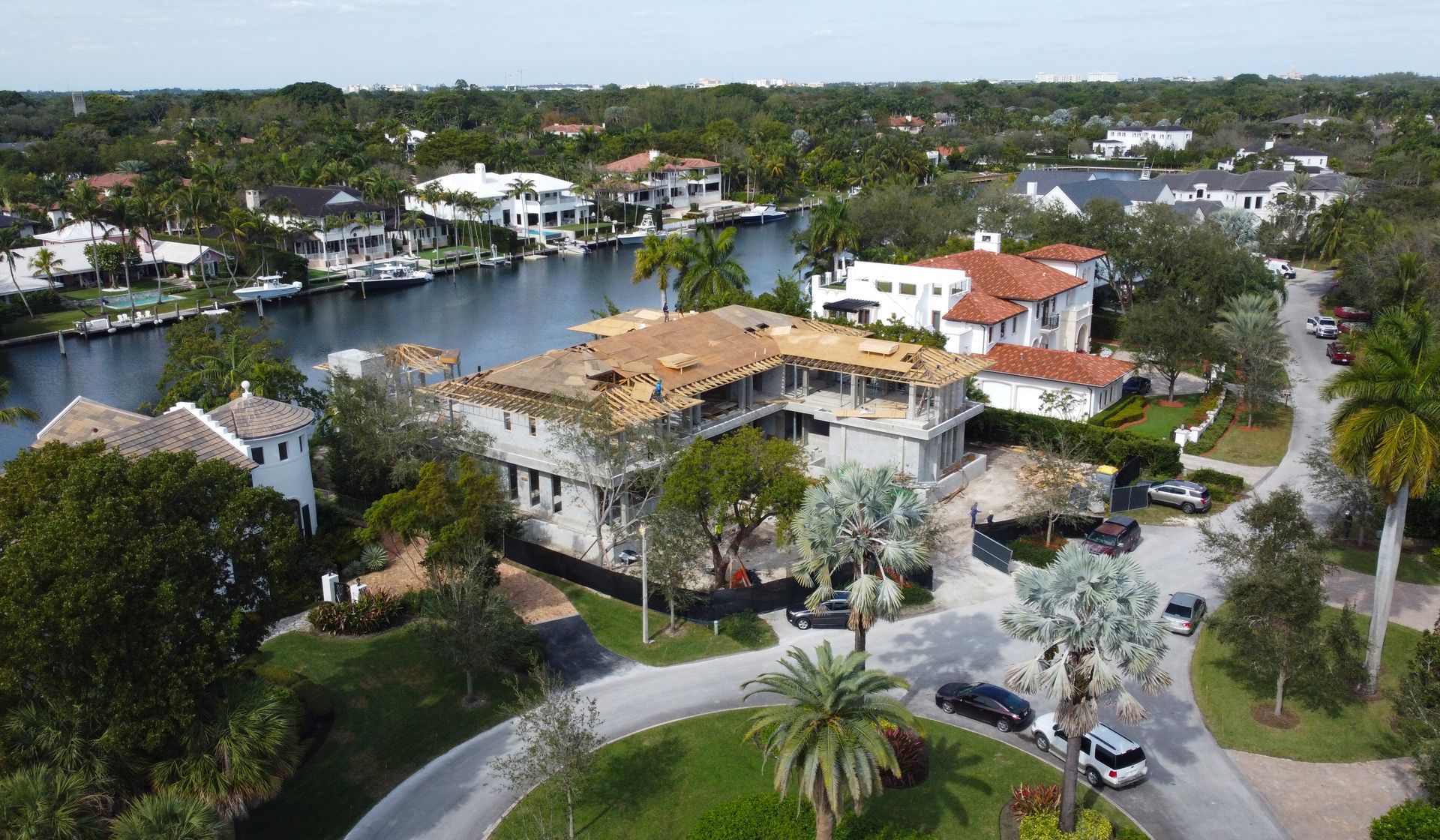 Aerial view of waterfront homes with boats and a large house under construction; trees and blue sky.