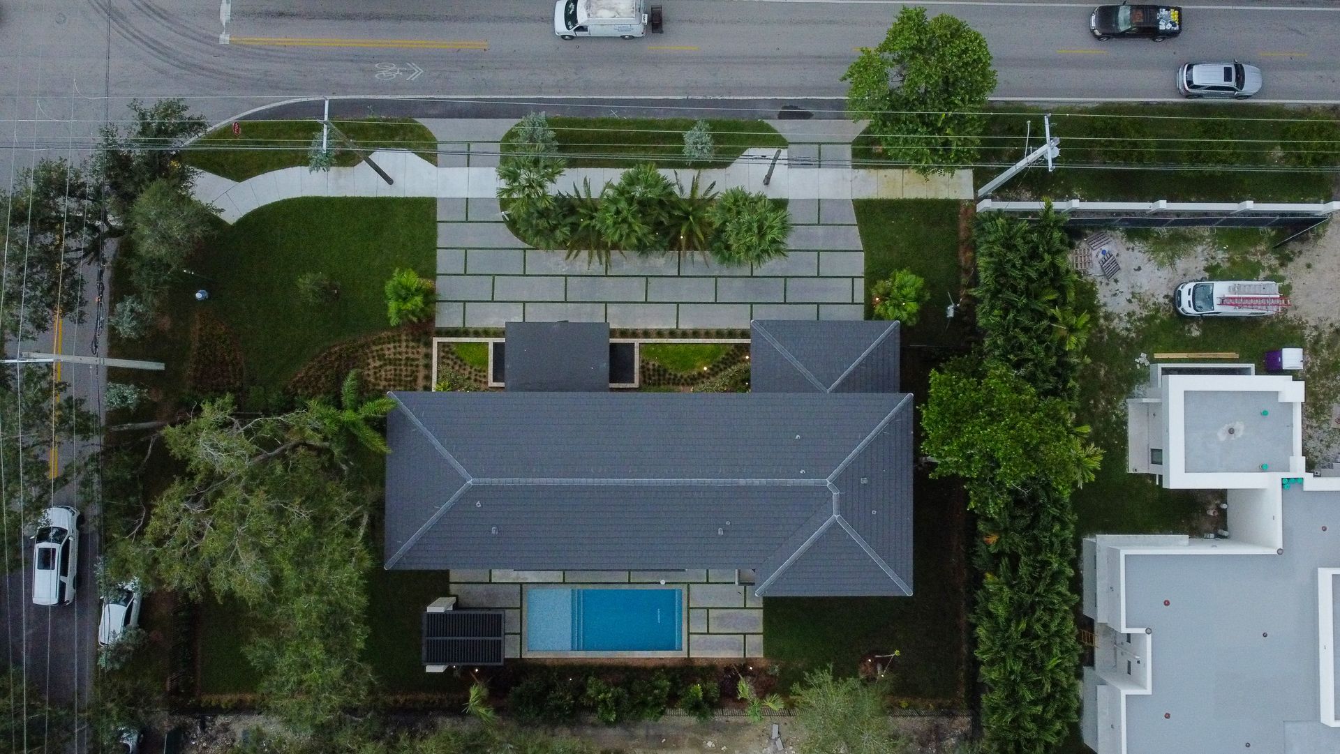 Aerial view of a home with a pool, gray roof, surrounded by green trees and grass, near a road.