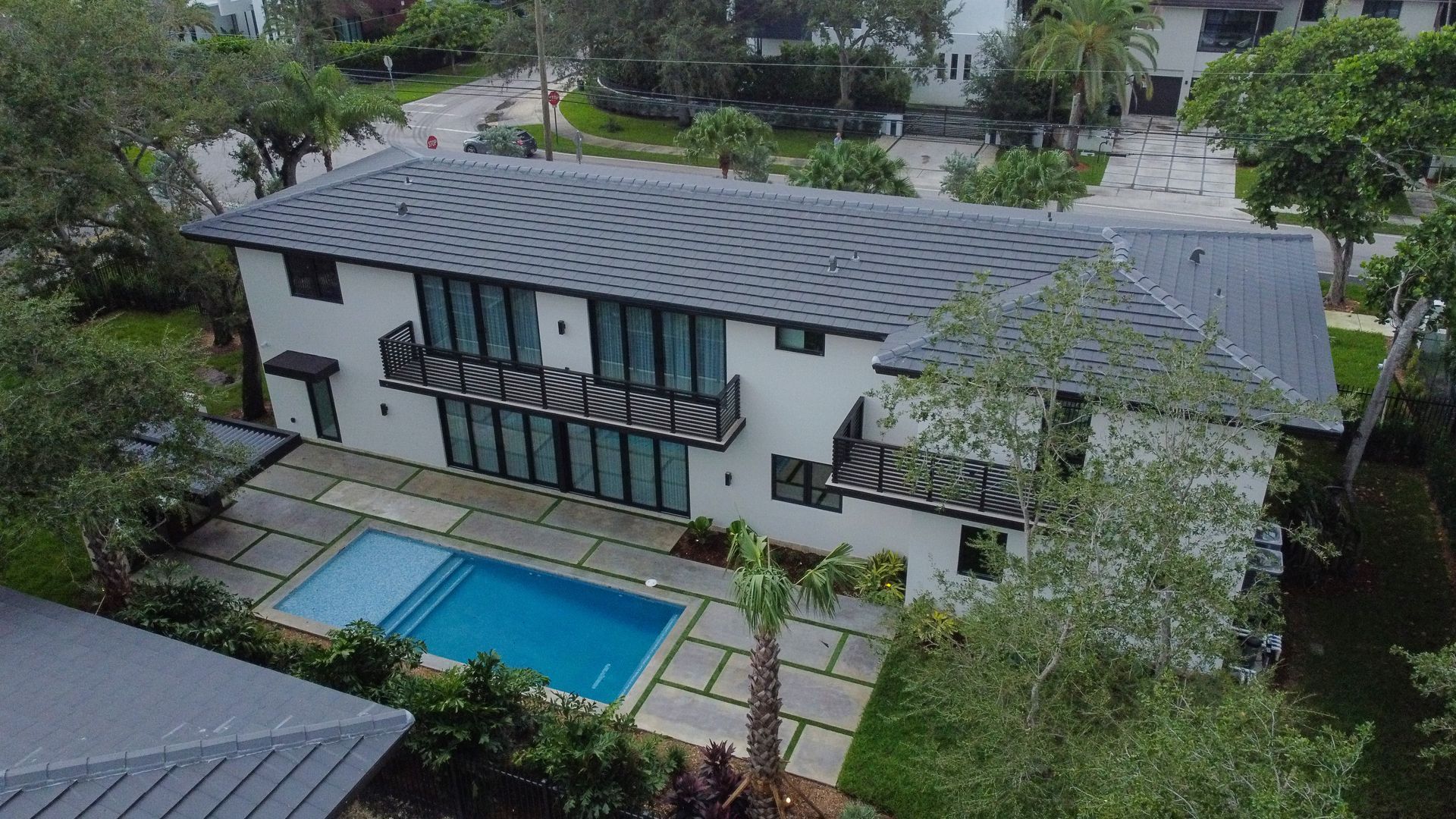 Modern two-story house with a pool. White walls, black trim, gray roof, surrounded by trees and landscaping.