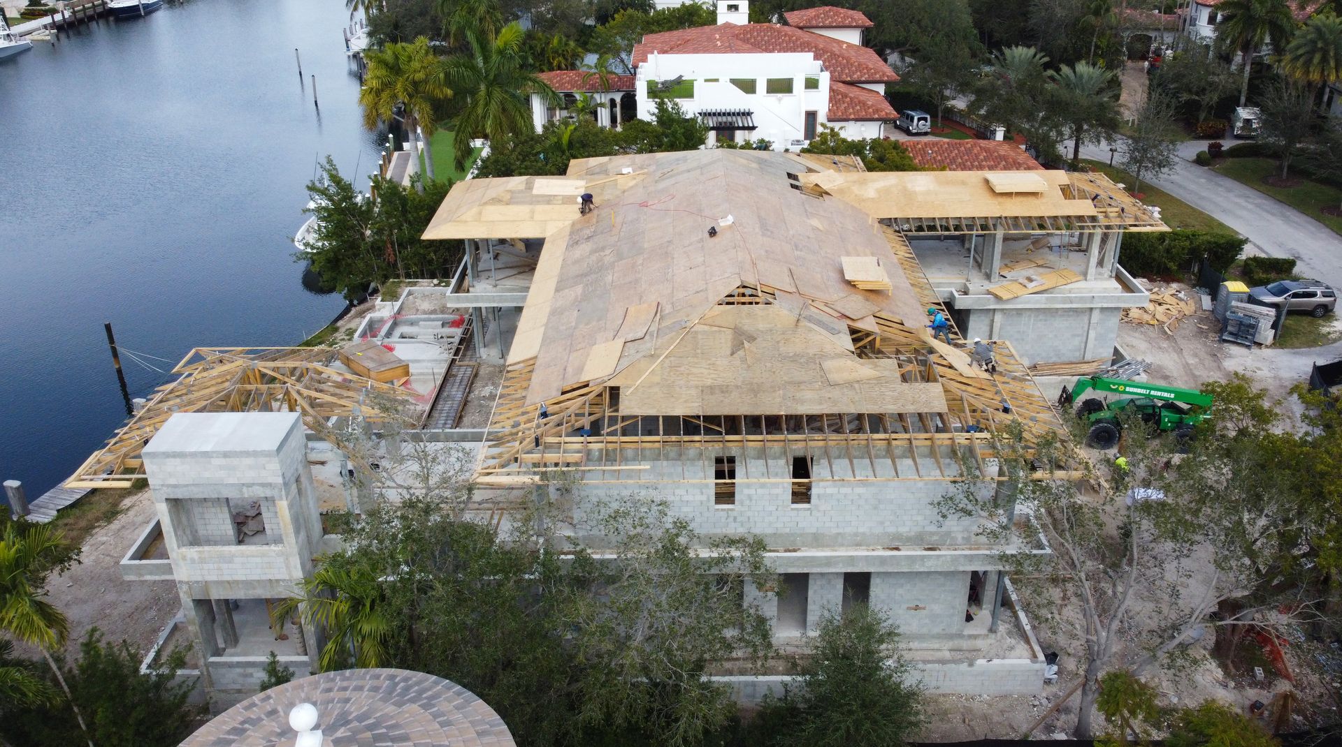 Construction site of a waterfront mansion; workers, unfinished roof, and materials visible.