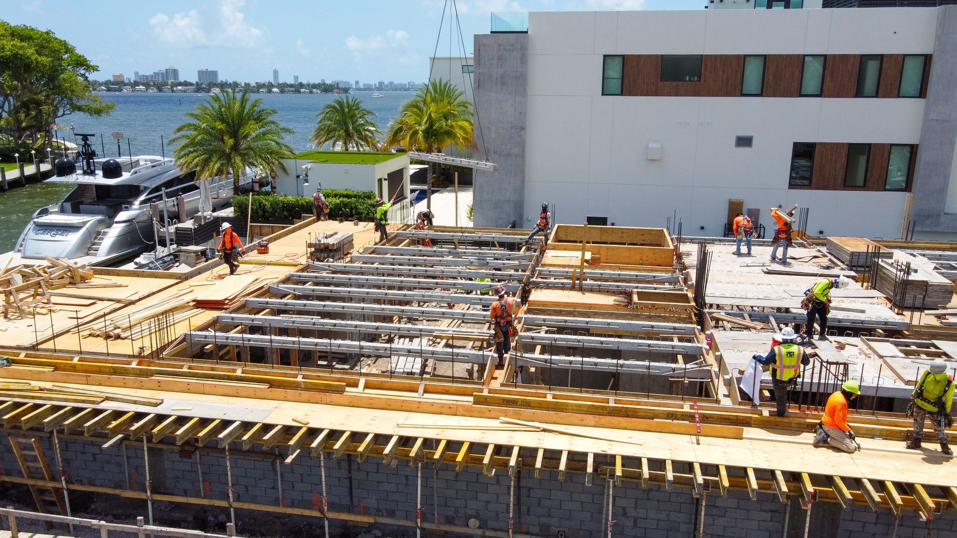 Construction site with workers, materials, and a yacht in the background. Building by water under a sunny sky.