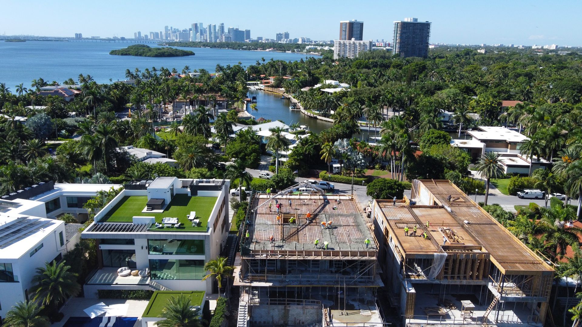 Aerial view of luxury homes; one under construction. City skyline and water visible in the background. Sunny day.