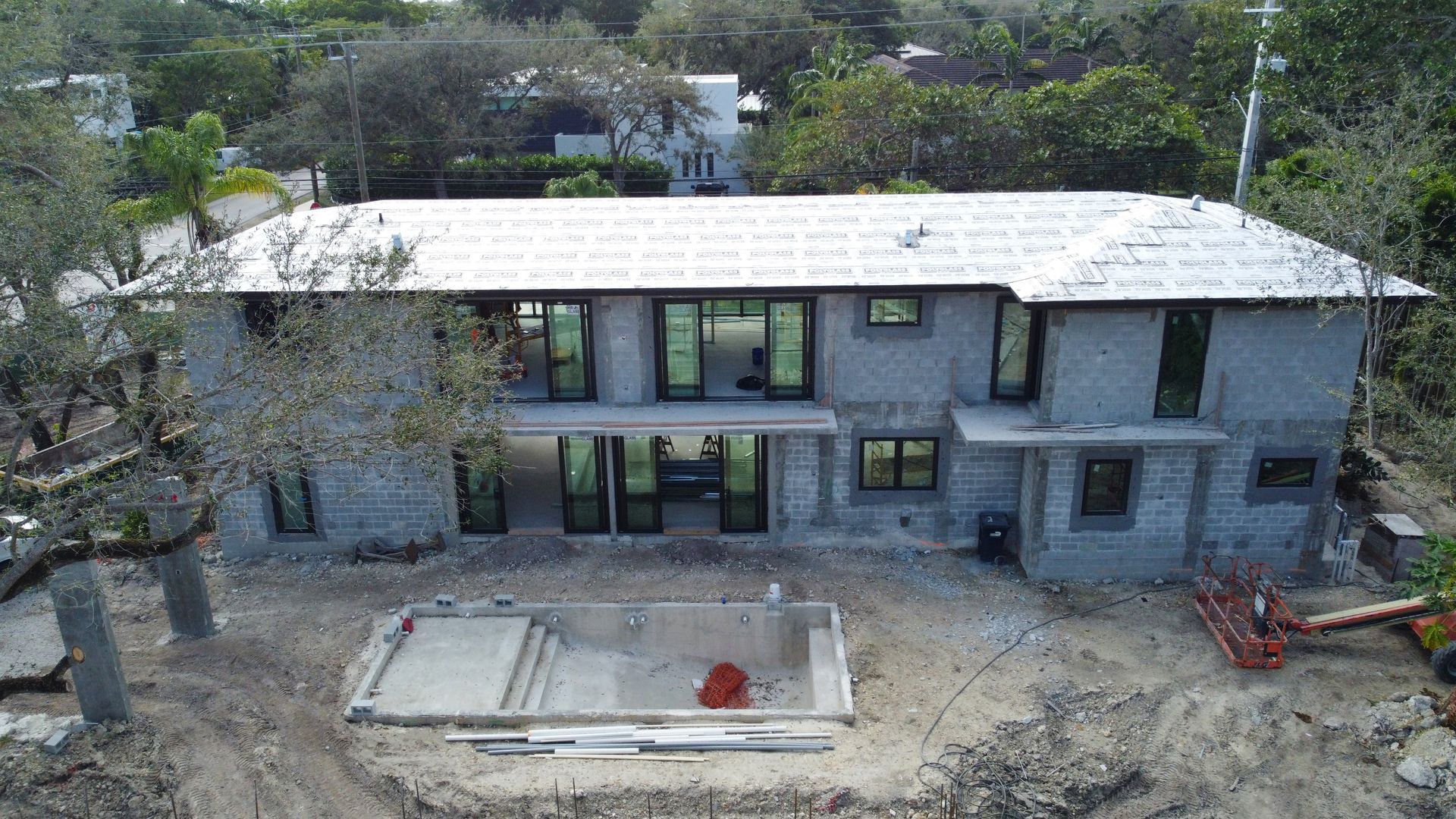 Two-story concrete house under construction with large windows and a rectangular pool foundation in front.
