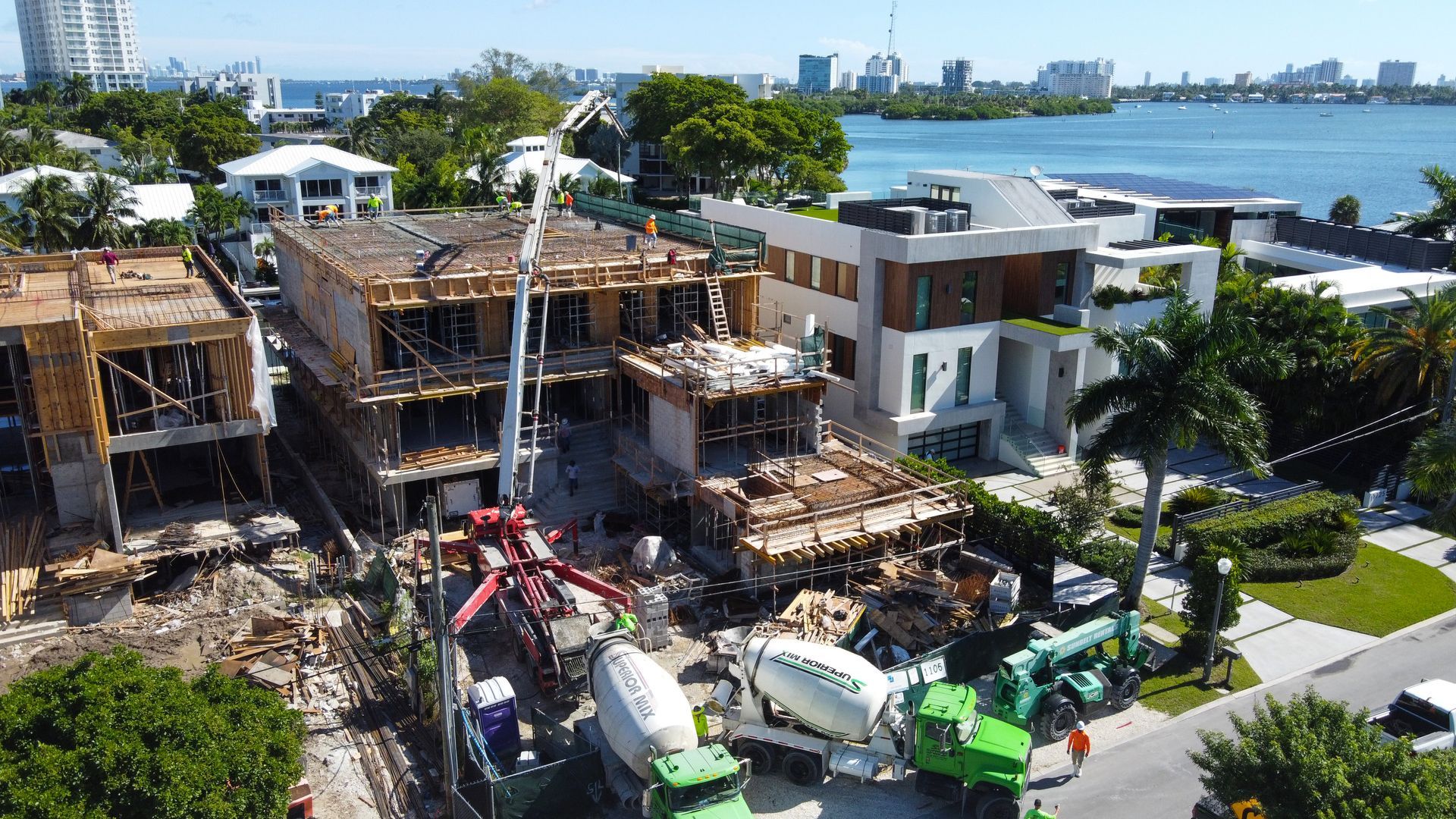 Construction site with multiple buildings, concrete mixers, and a crane. Bright, sunny day, waterfront view.