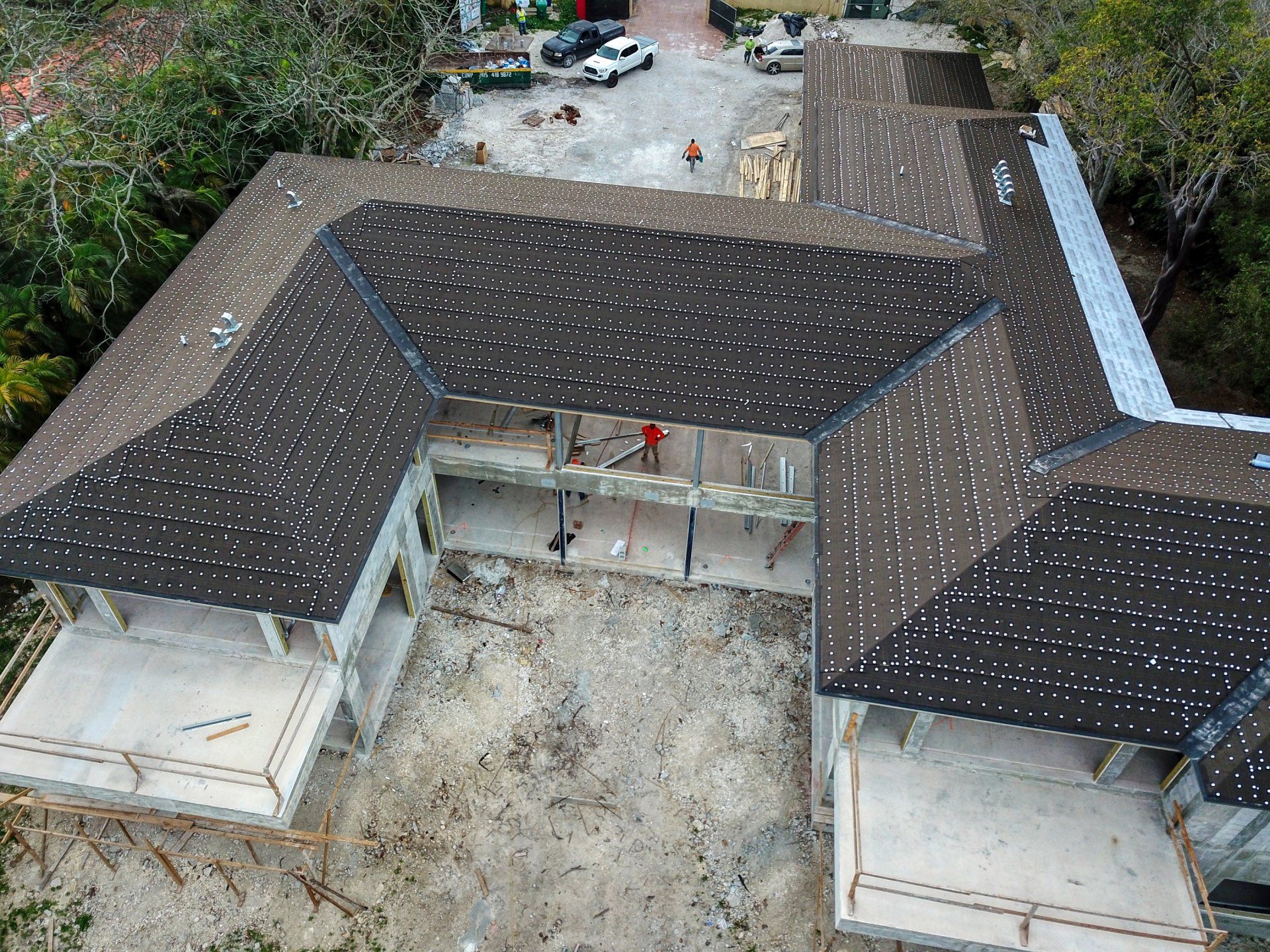 Aerial view of a building under construction, dark roof with white speckles, surrounded by debris and construction equipment.