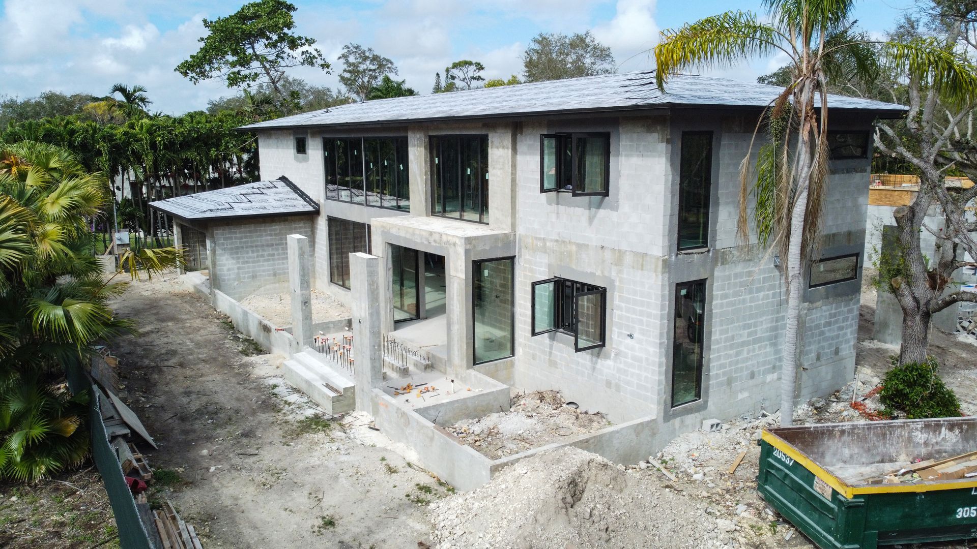 Two-story house under construction, with gray stone exterior and large windows. Construction debris visible around the foundation.