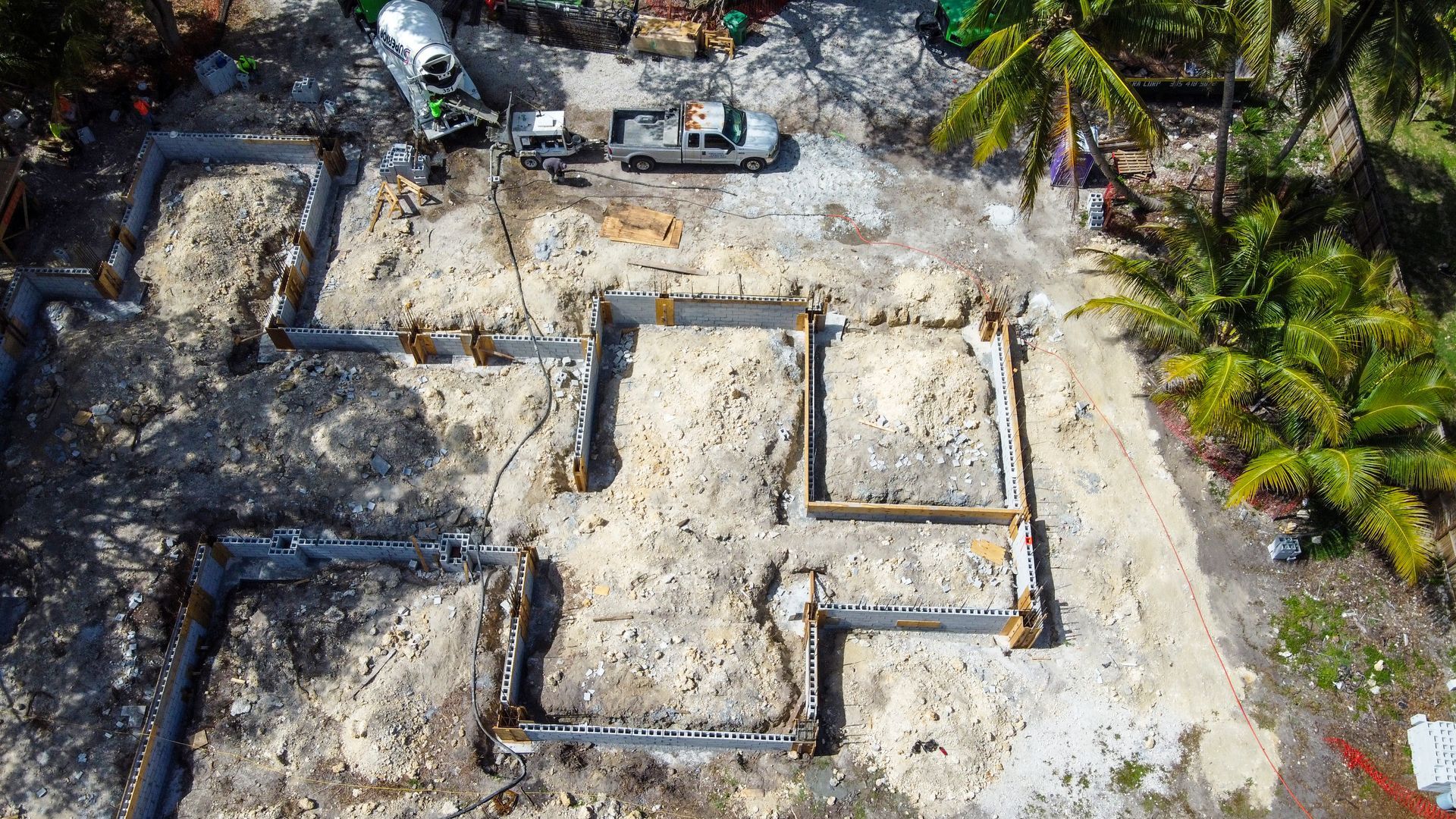 Construction site: foundation outline with dirt and equipment, including a cement truck, in a tropical setting.
