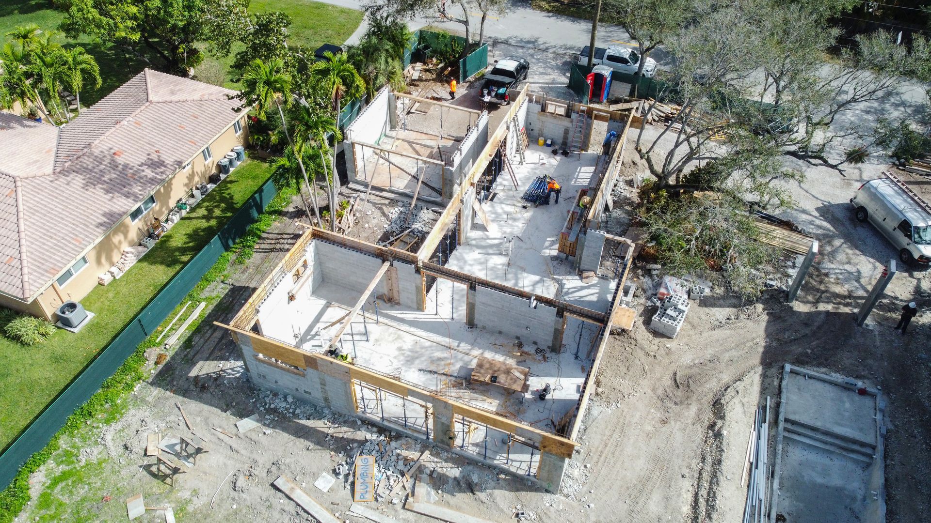 Construction site: foundation, wood framing, workers, vehicles, nearby houses, trees.