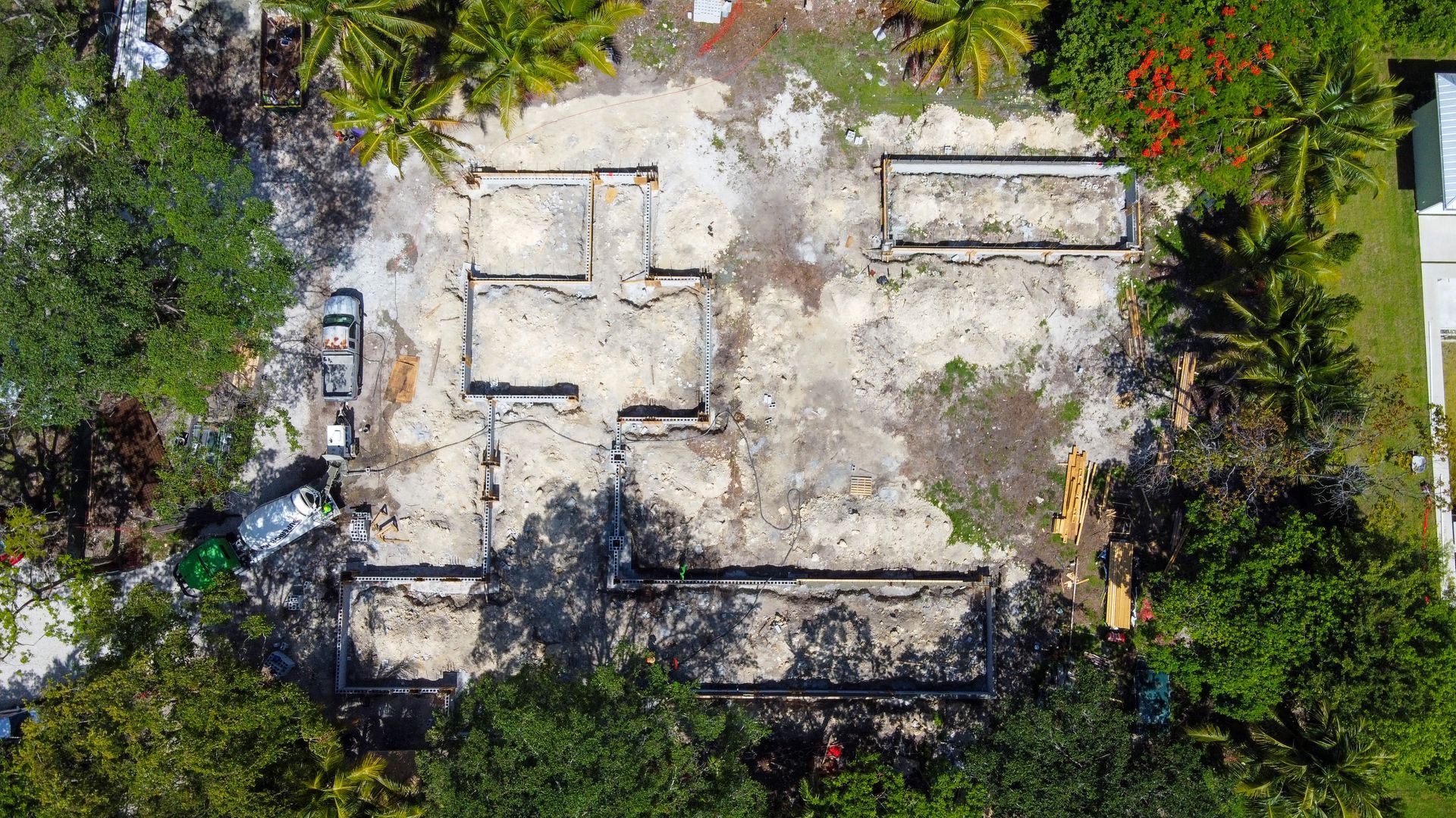 Aerial view of a construction site. Concrete foundation outline amid dirt, with surrounding trees and foliage.