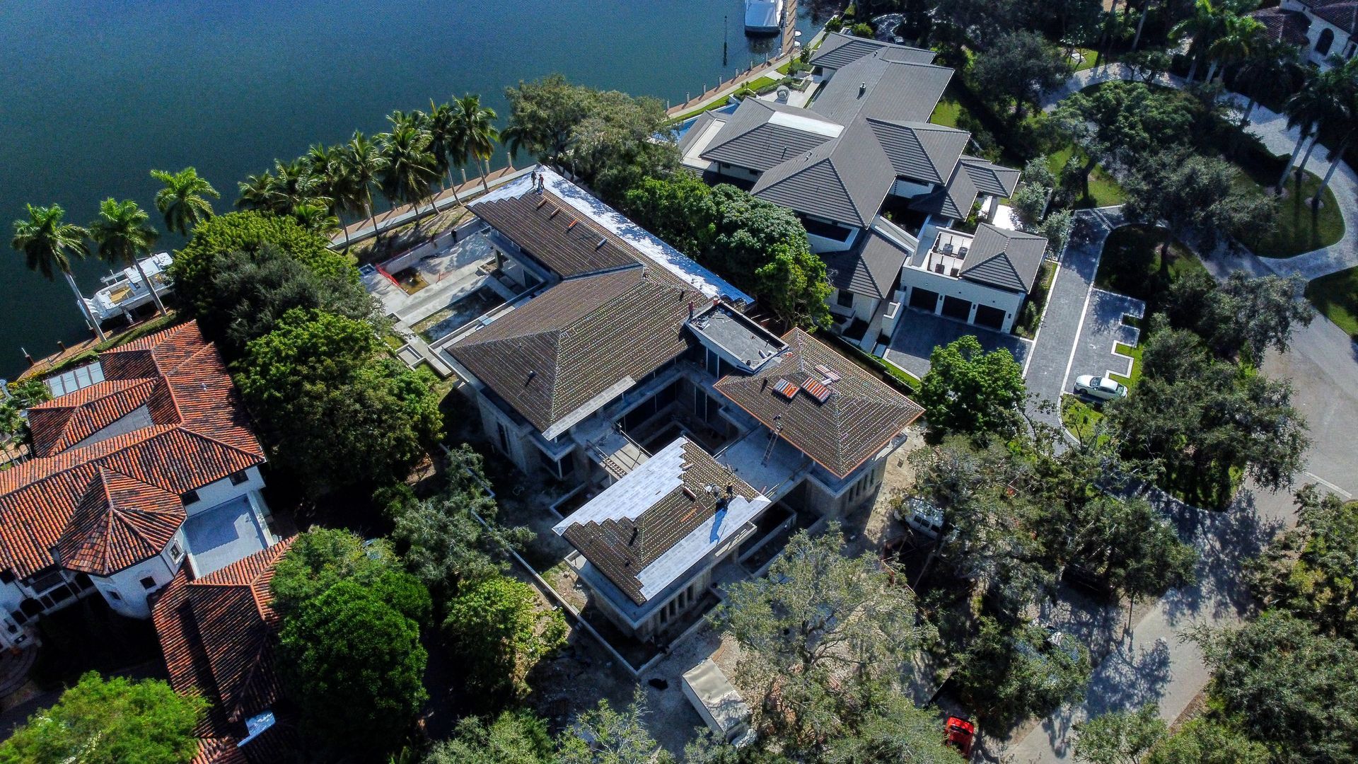 Aerial view of a large house with a dark roof surrounded by trees near a body of water.