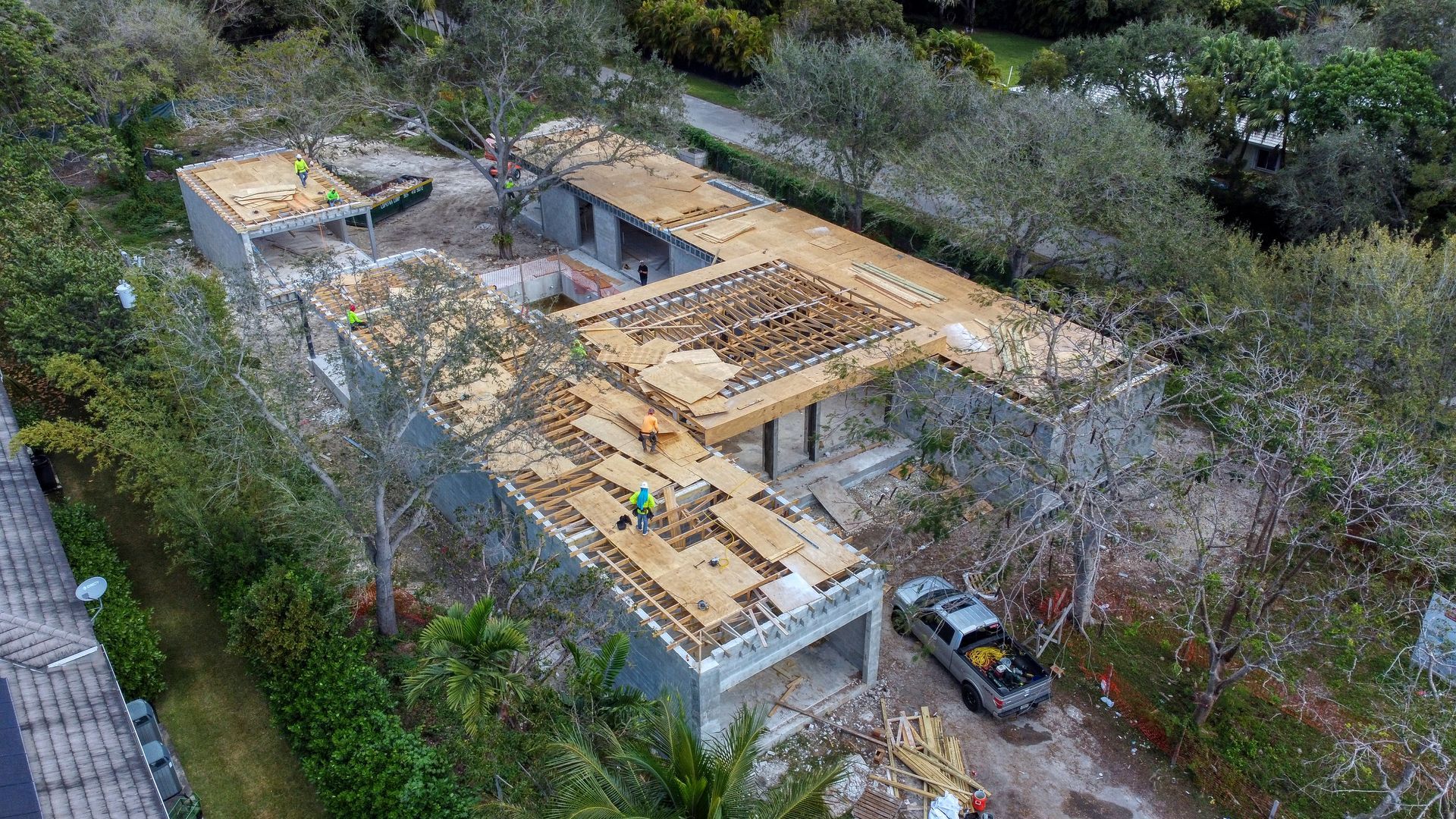 Construction site: workers on roofs of a U-shaped building, surrounded by trees, with a truck parked nearby.