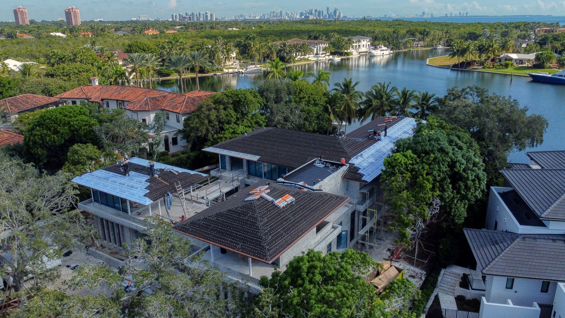 Aerial view of waterfront houses with clay tile roofs, lush green trees, and a distant city skyline.