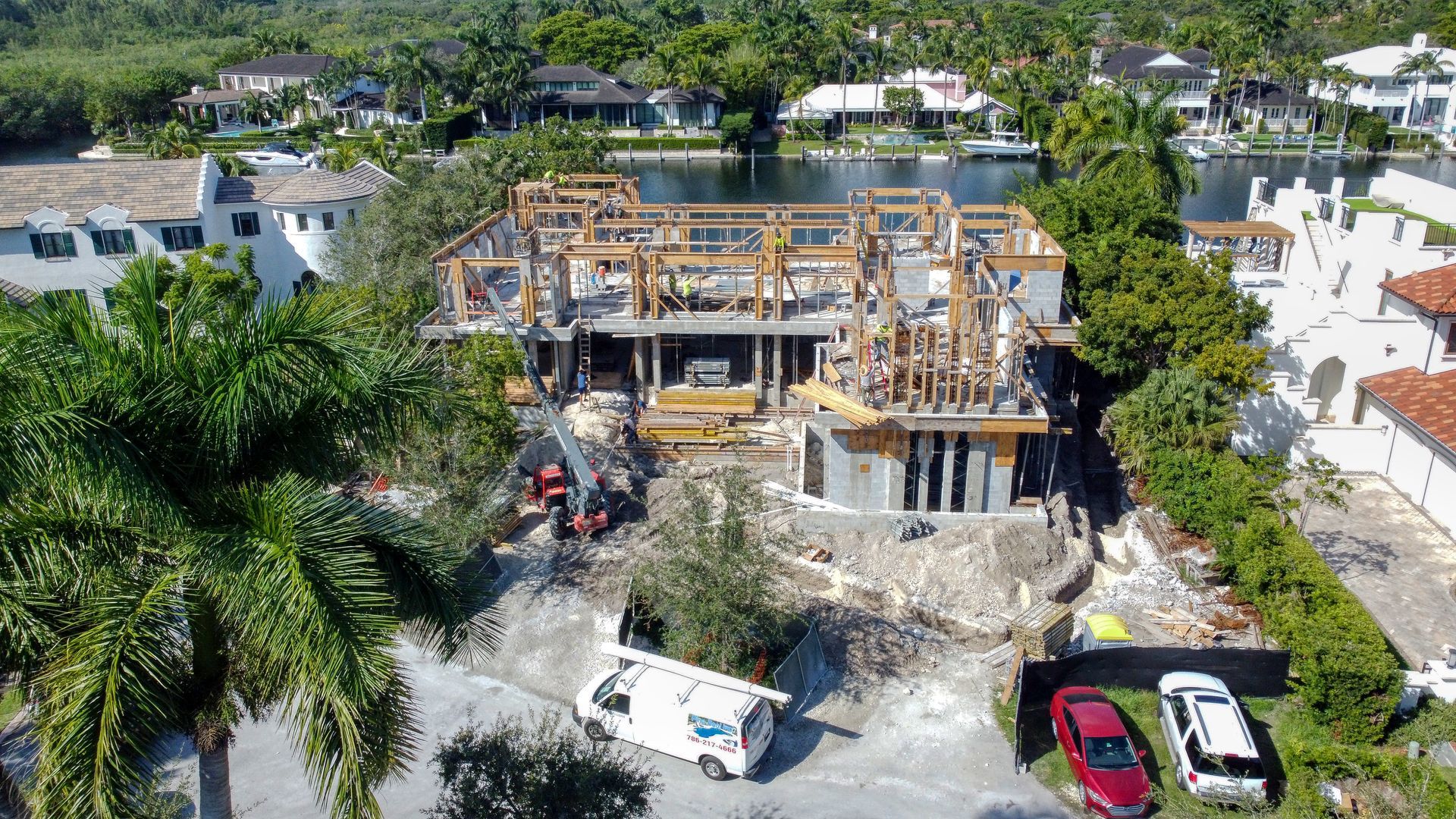 Construction site of a multi-story home with exposed wood framing, white van, and parked cars near water.