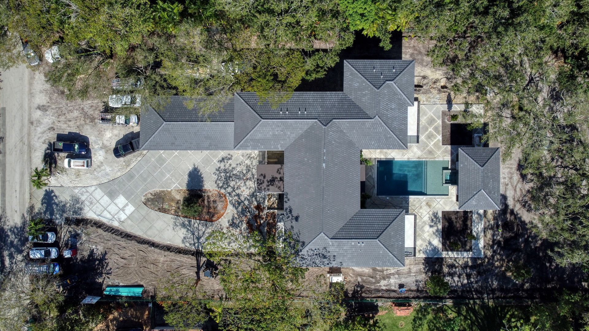 Aerial view of a gray-roofed house with a pool, surrounded by trees and a driveway.