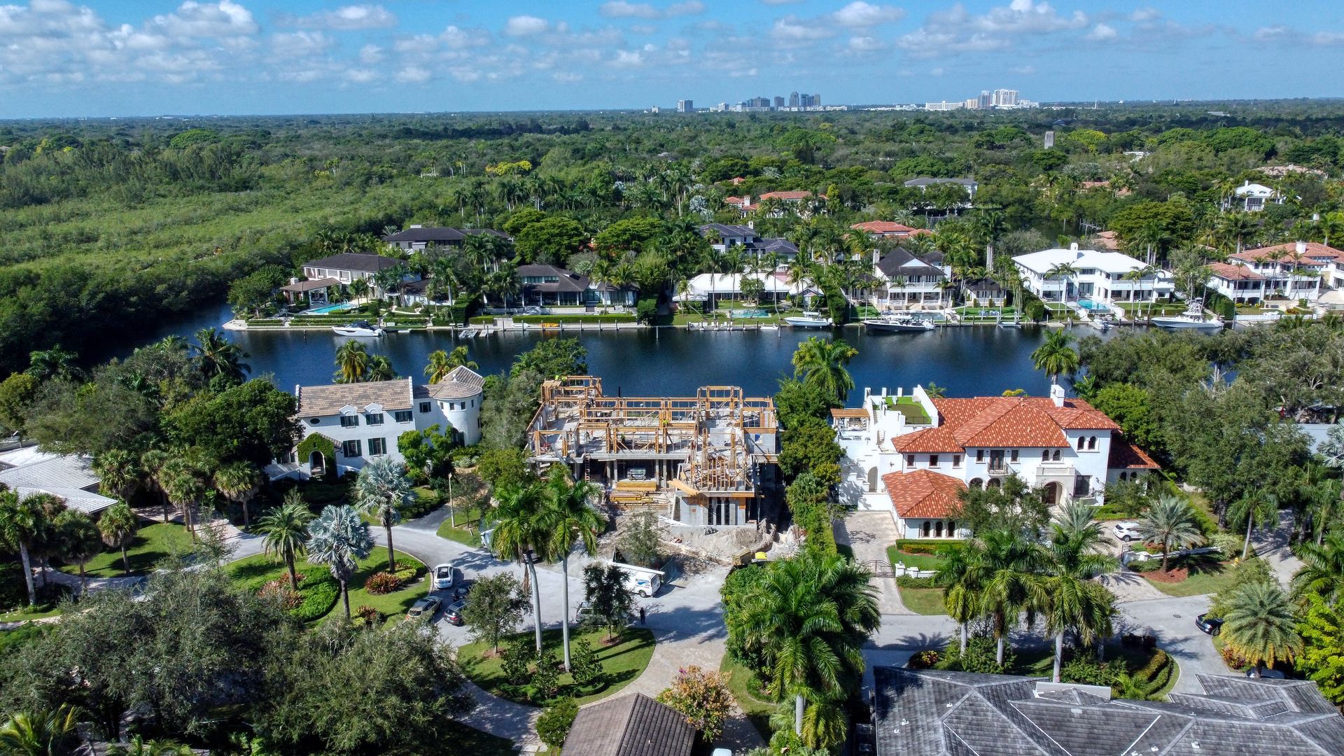 Aerial view of luxury homes on a waterway lined with lush greenery; city skyline in the distance.