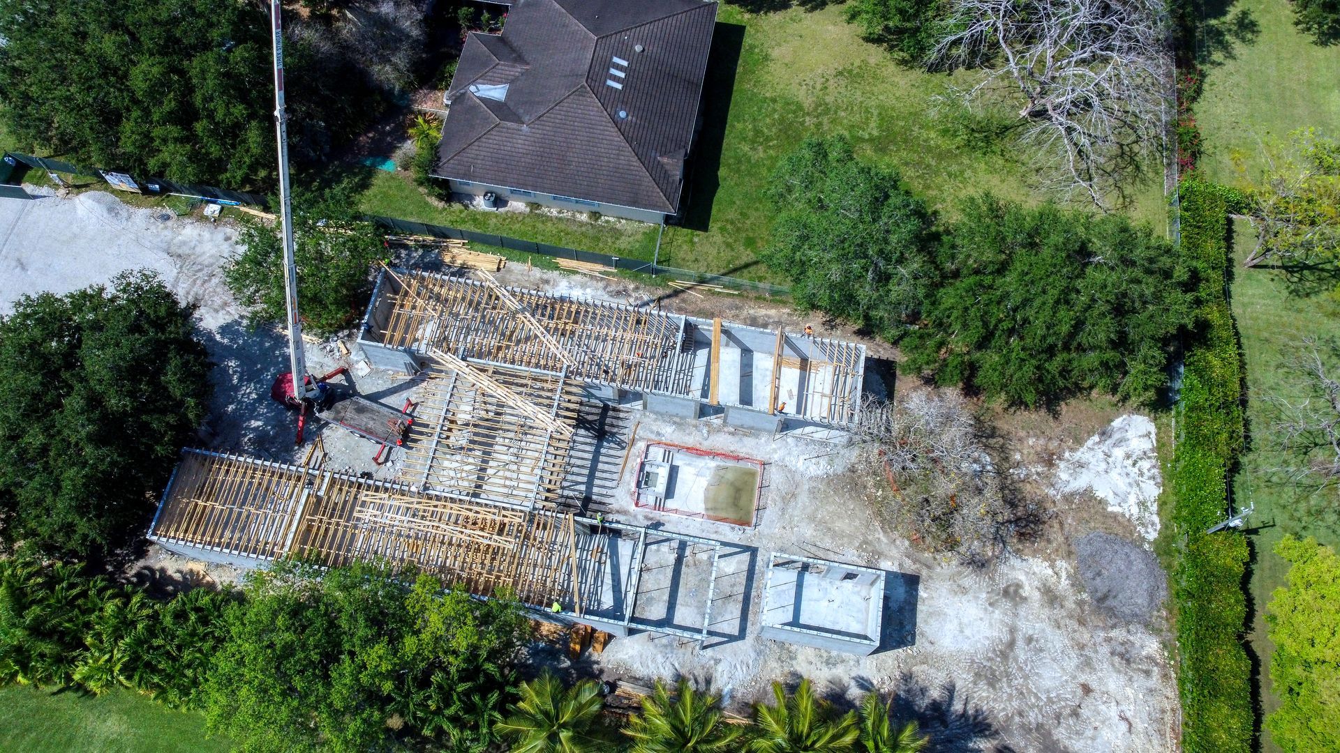 Aerial view of a construction site with building foundations, surrounded by trees and a nearby house.