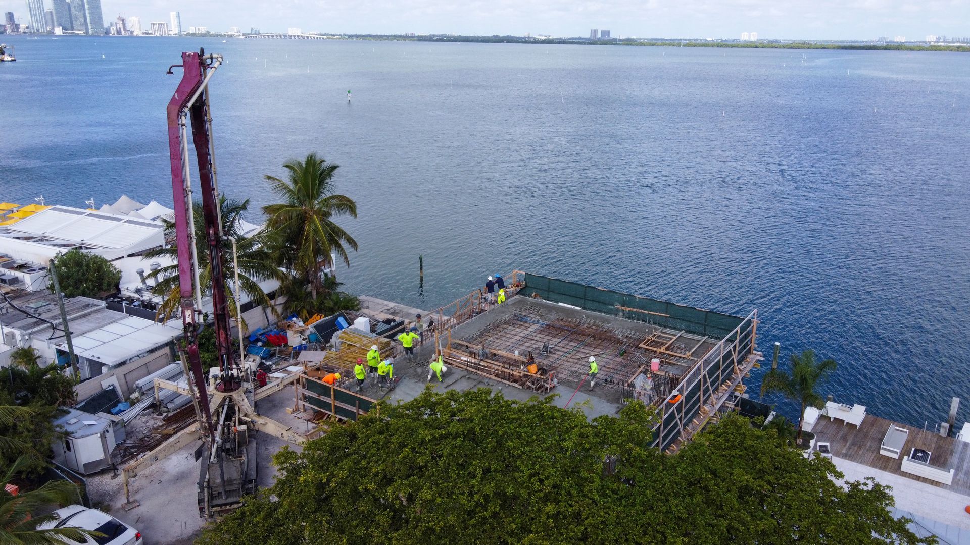 Construction site near water with a concrete pump pouring cement onto a building's roof; workers in yellow vests.