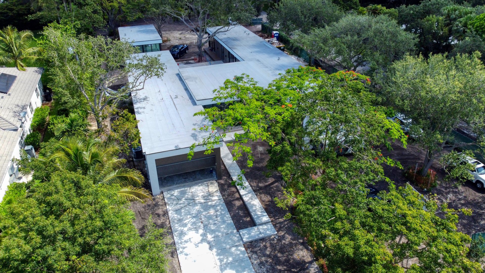 Aerial view of a modern house with a flat roof, surrounded by lush green trees and a concrete driveway.