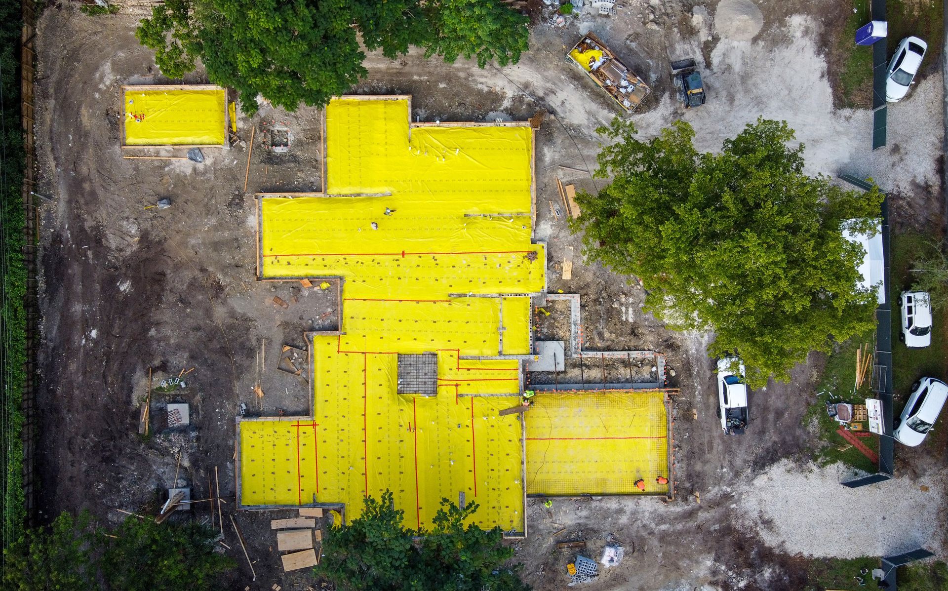 Overhead view of a building under construction, yellow panels laid out on foundation; equipment and trees surround the site.