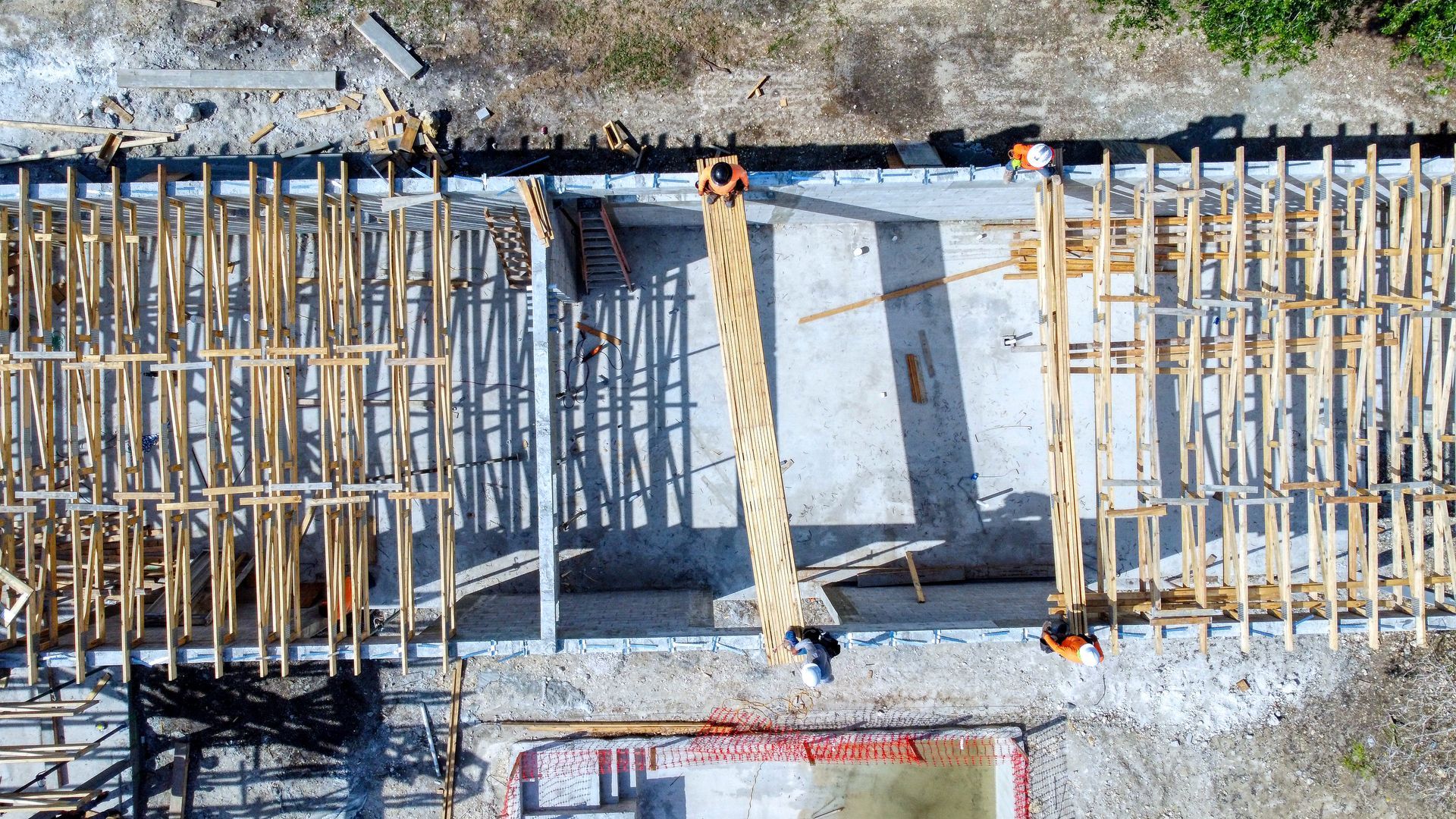 Overhead view of a building under construction, wooden beams laid out on concrete. Workers are visible on site.