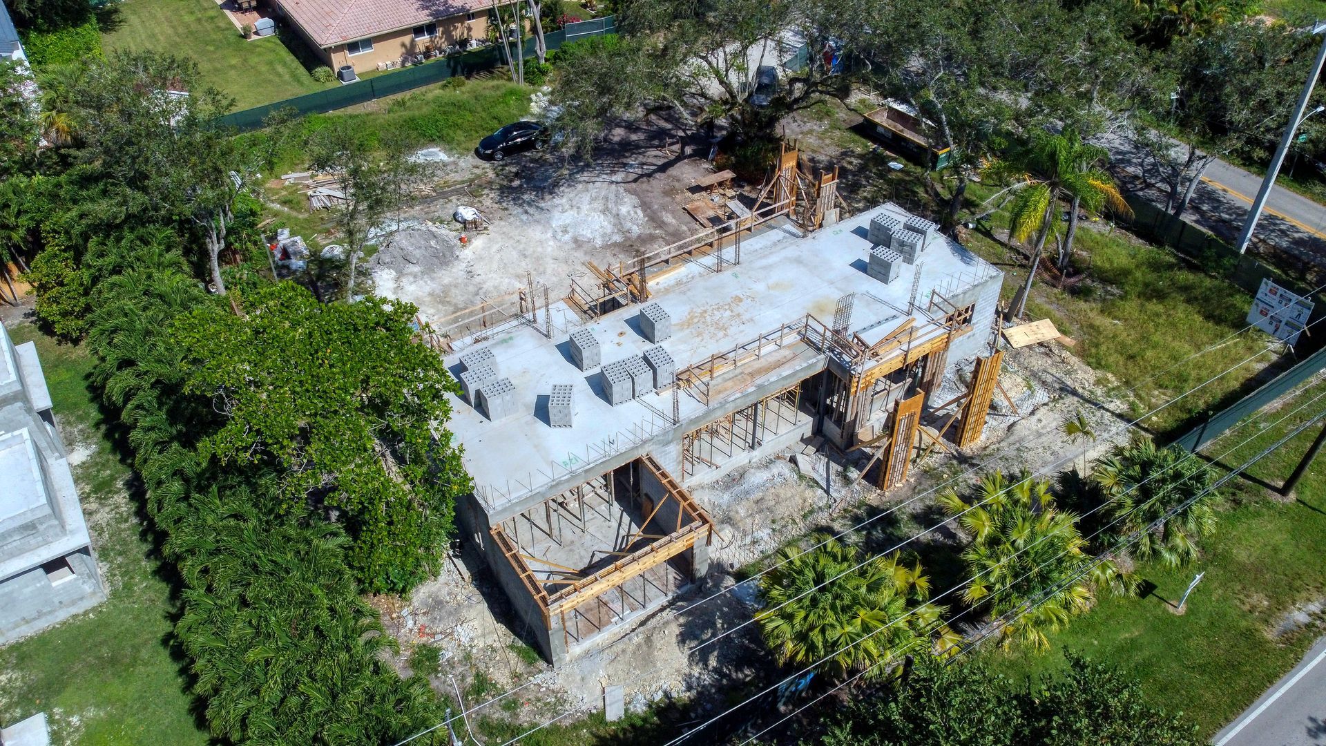 Aerial view of a building under construction, showing a concrete foundation and exposed framing, surrounded by trees and a green fence.
