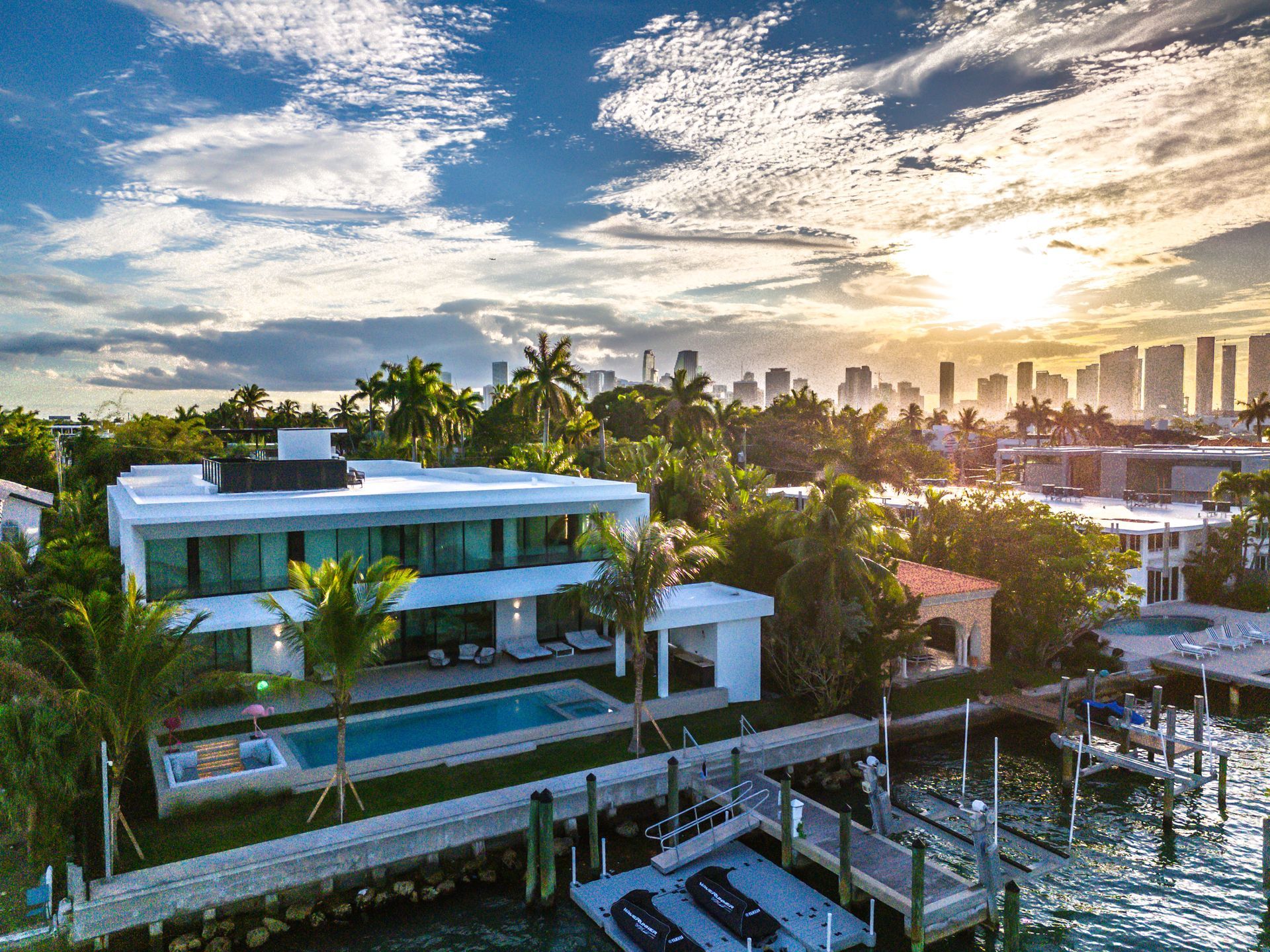 Modern waterfront home with pool and dock at sunset, Miami skyline in the background.