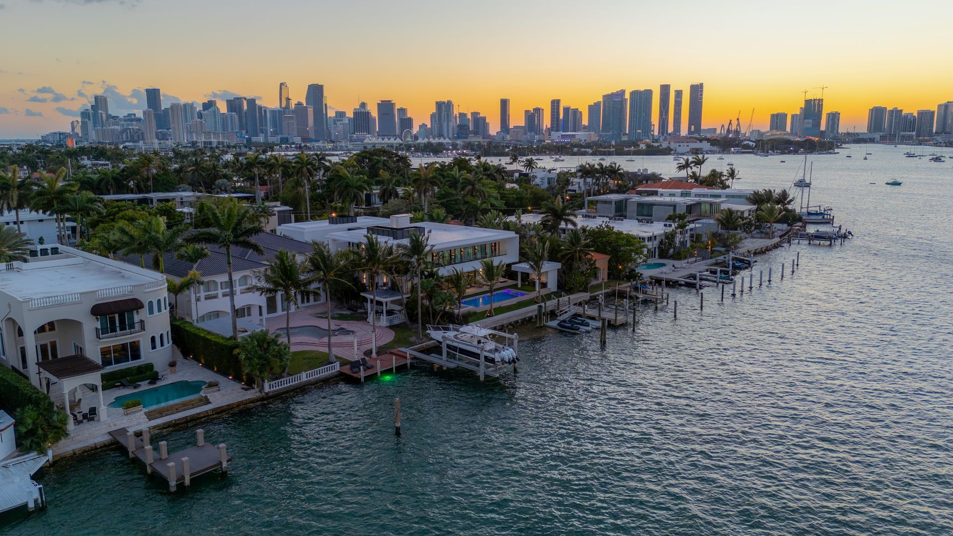 Waterfront homes at sunset, city skyline in the background. Calm water, docks, and lush greenery.