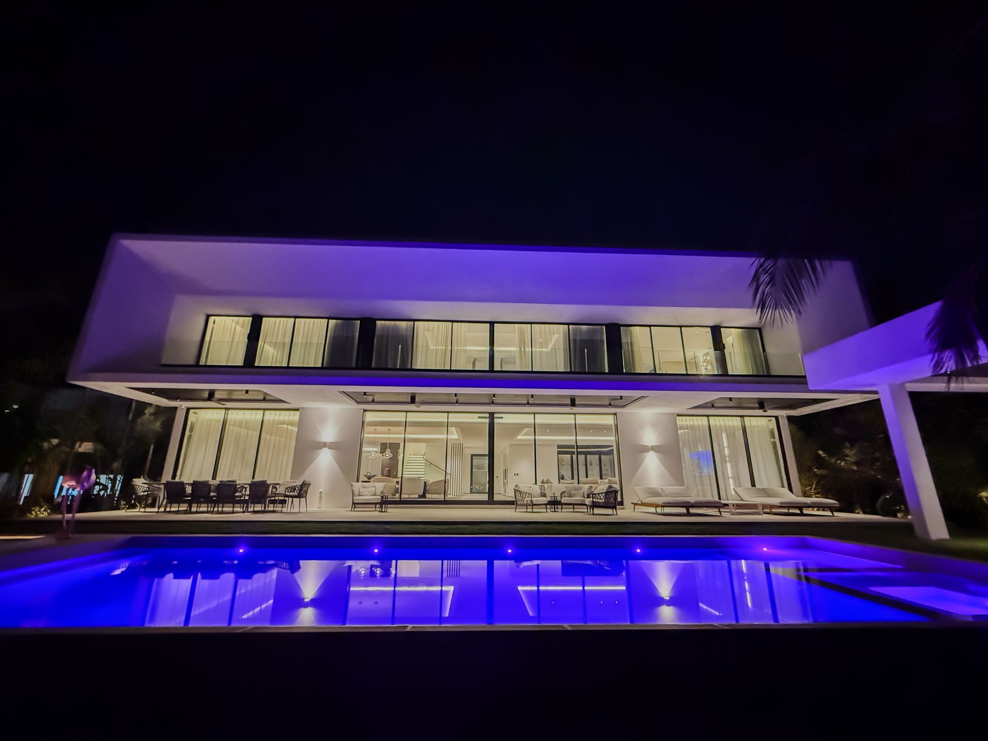 Modern white house at night with a blue-lit pool. Large windows reveal interior lights.