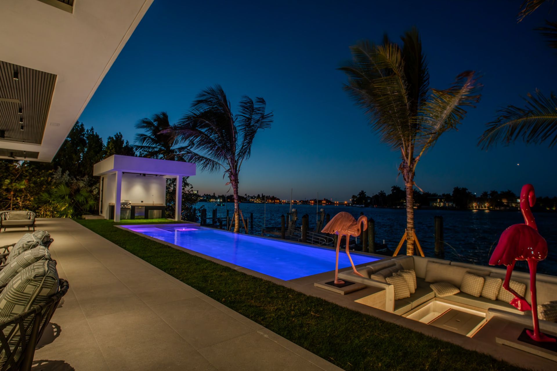 Night view of a waterfront home with pool, illuminated in blue, with pink flamingo statues and a gazebo.