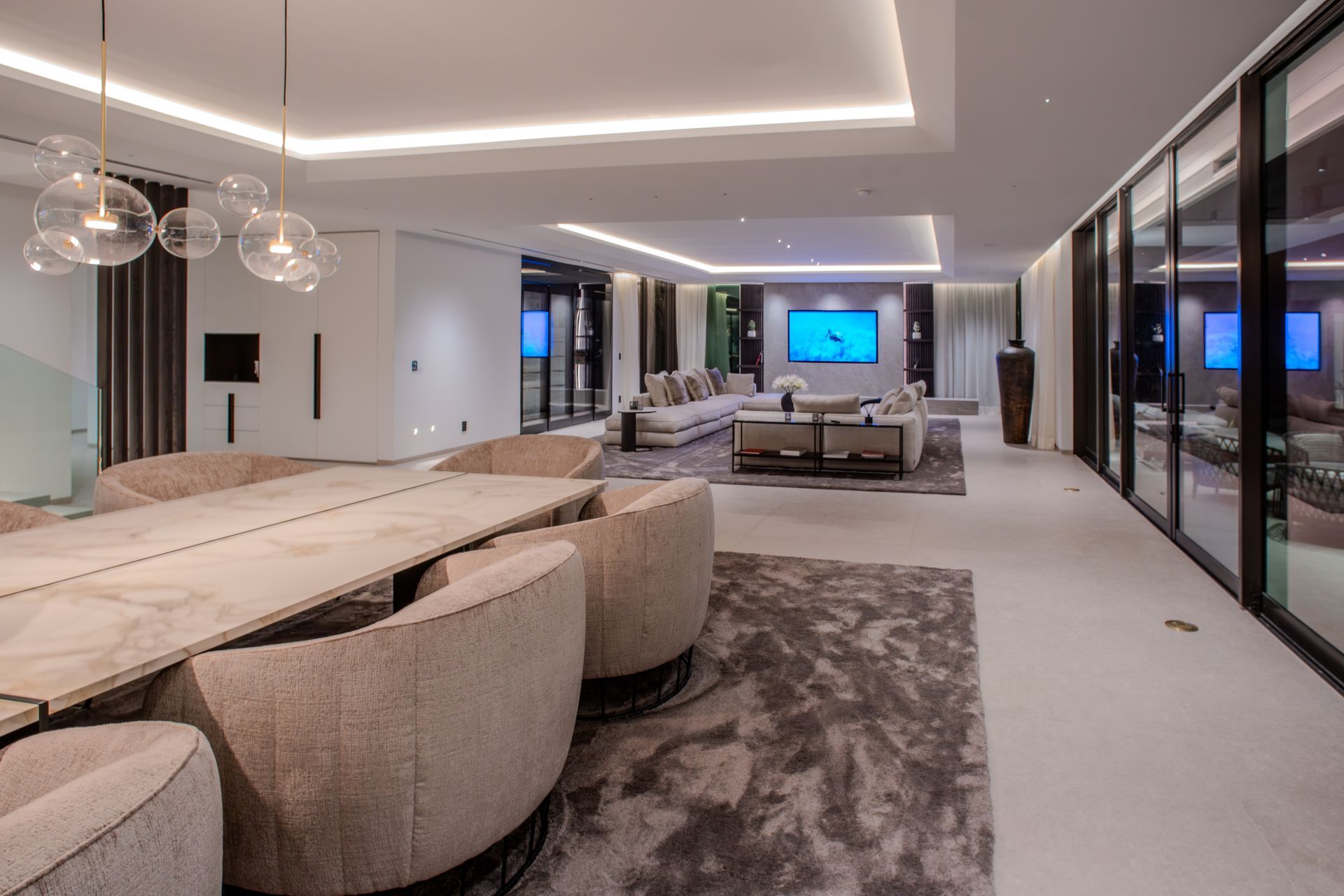 Modern, open-concept living space with dining table, sofa, and floor-to-ceiling windows. Neutral tones and ambient lighting.