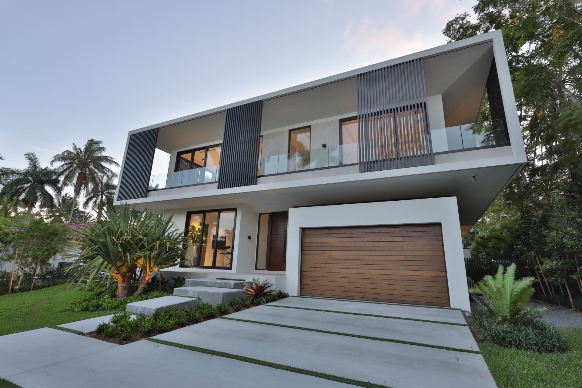 Modern two-story house with a wooden garage door and glass balconies, set on a green lawn with palm trees.