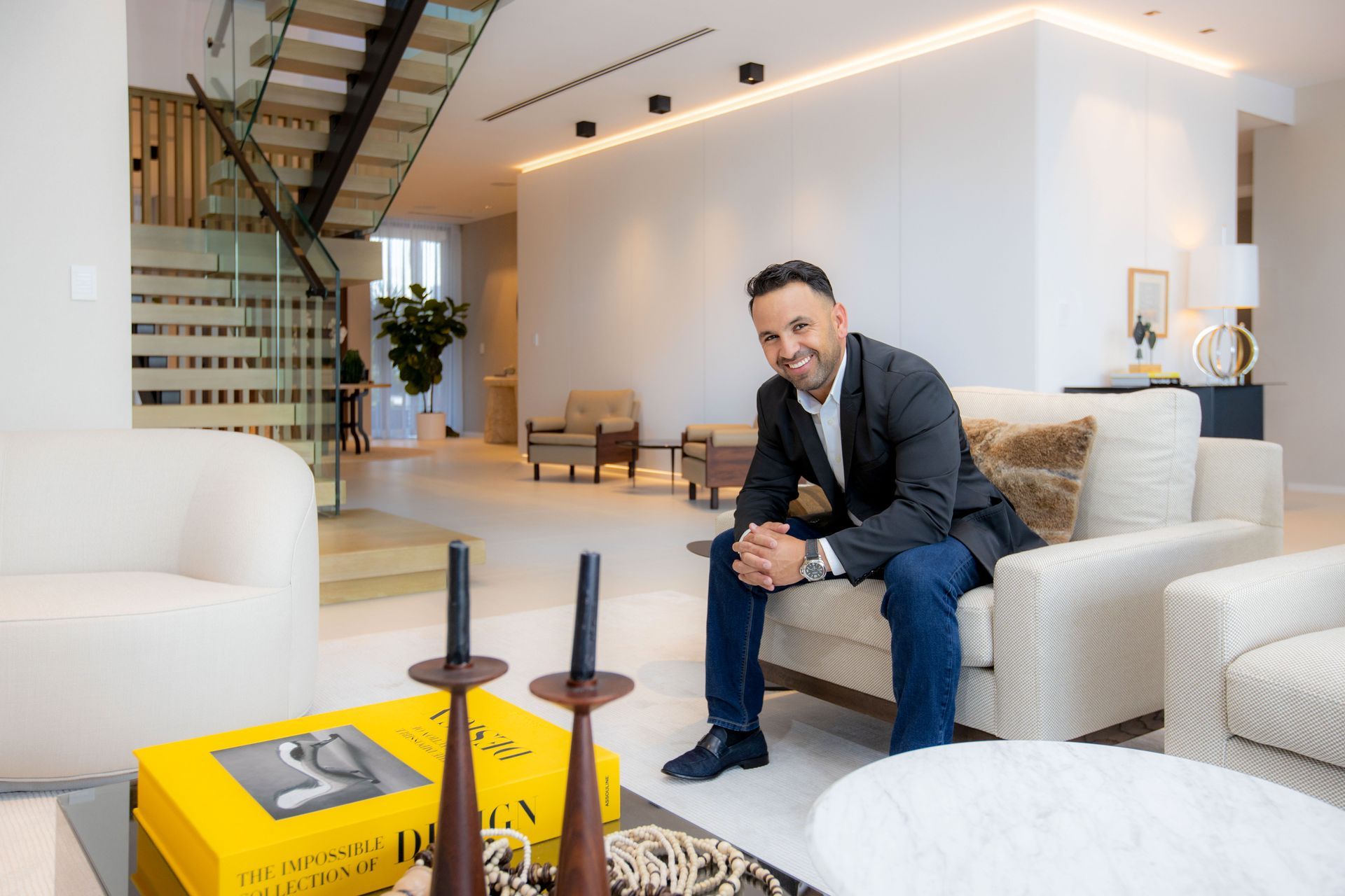 Man sitting on a sofa in a modern living room, smiling. Yellow book, wood accents.
