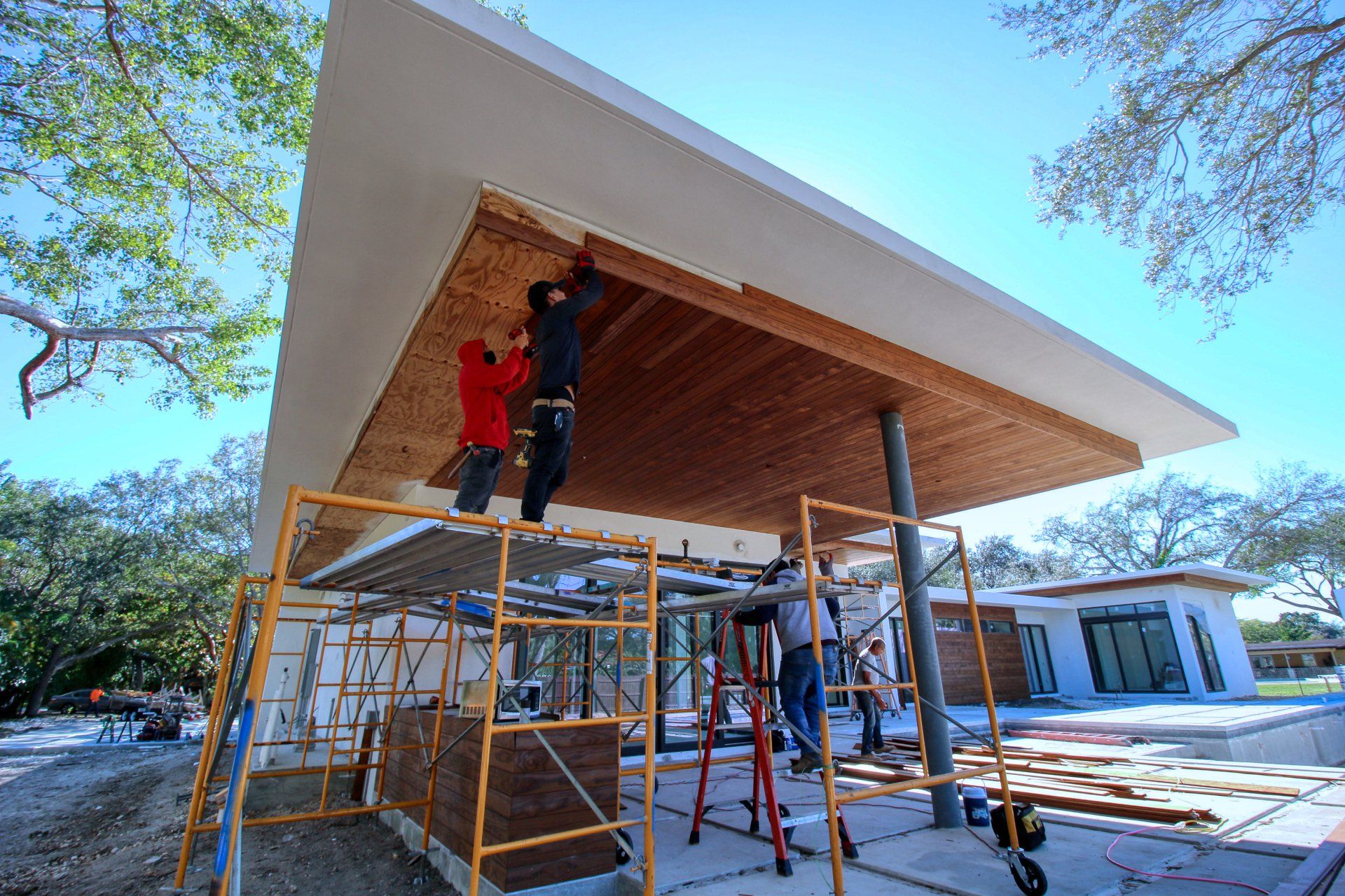 Construction workers installing wood paneling on the underside of a modern home's roof. Scaffold and materials visible.