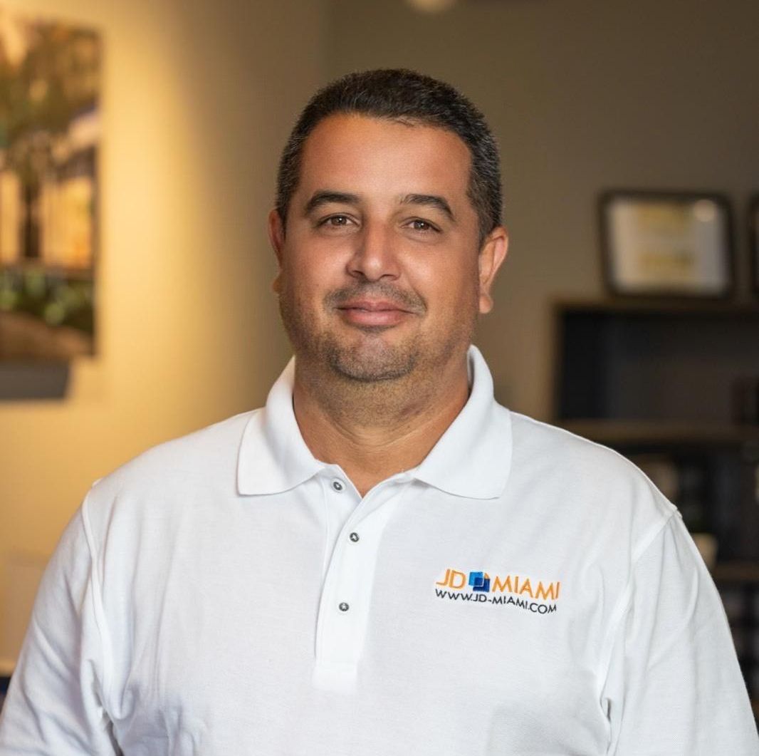 Man in white polo shirt, smiling, in an office setting with a logo on the shirt.
