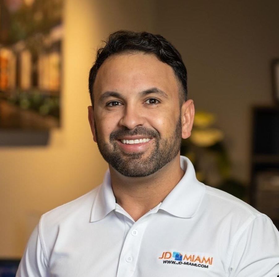 Man smiling, wearing a white polo shirt, logo on shirt, in an office setting.
