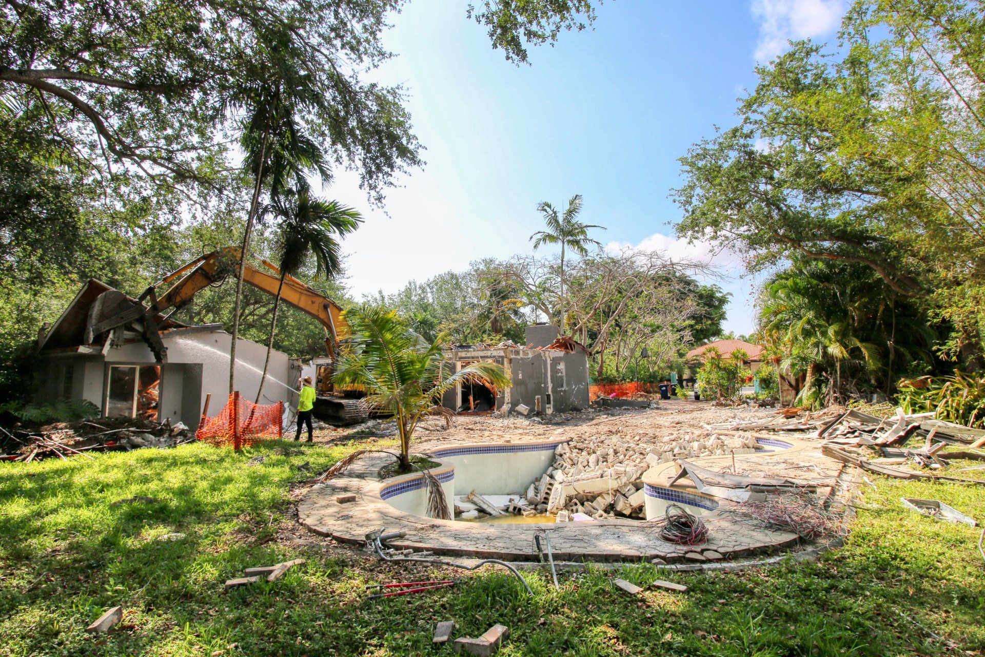 Demolition of a house and pool, excavator removing debris. Green lawn, trees, blue sky.