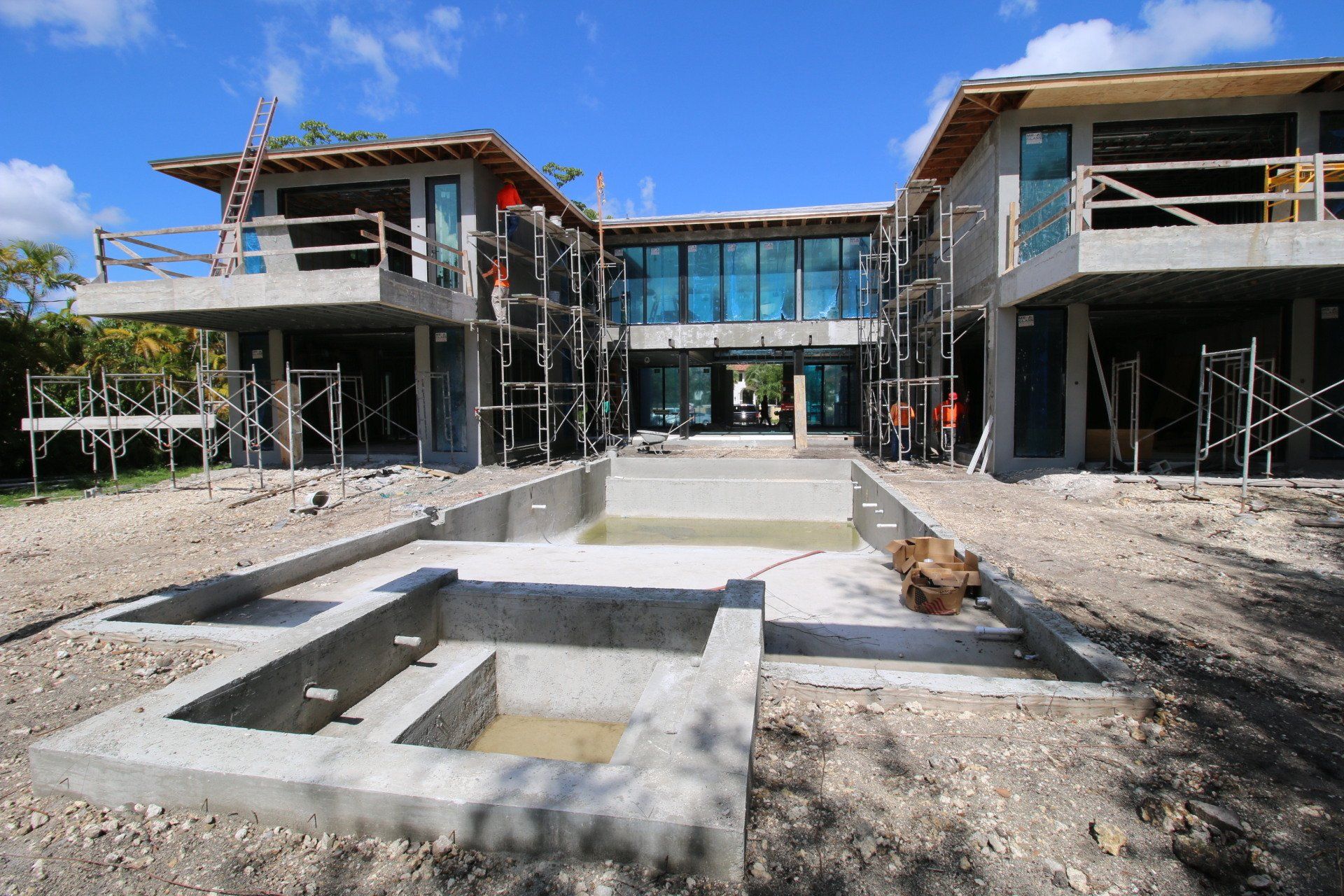 A house under construction with concrete walls, a pool, and scaffolding on a bright, sunny day.