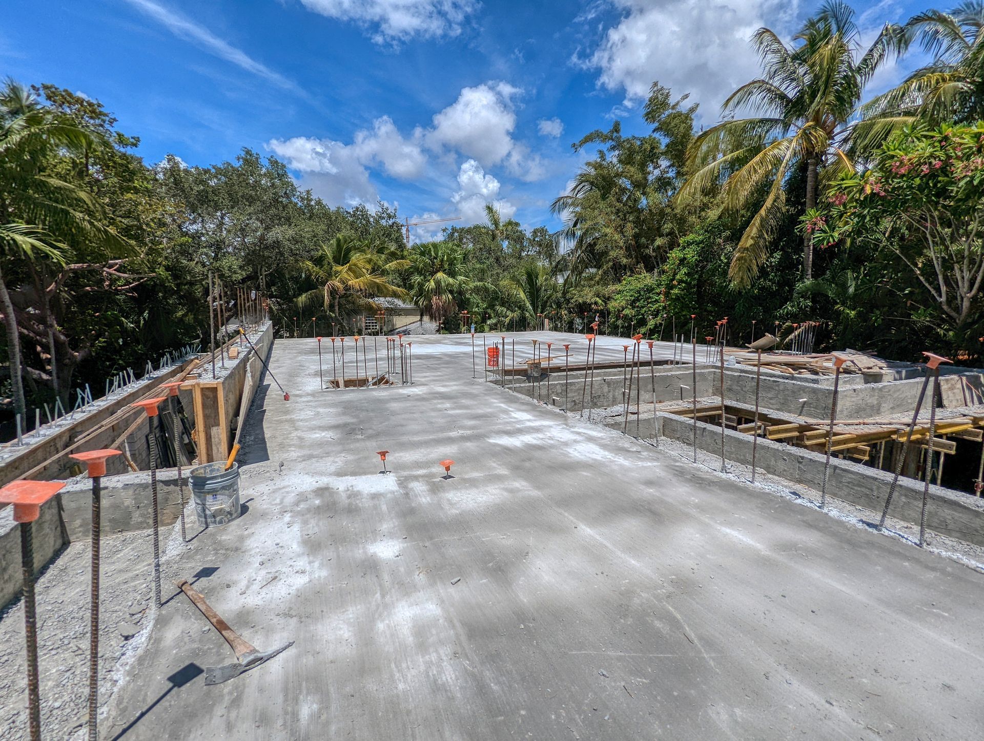 Concrete construction site with gray surfaces, surrounded by trees and a blue sky with clouds.