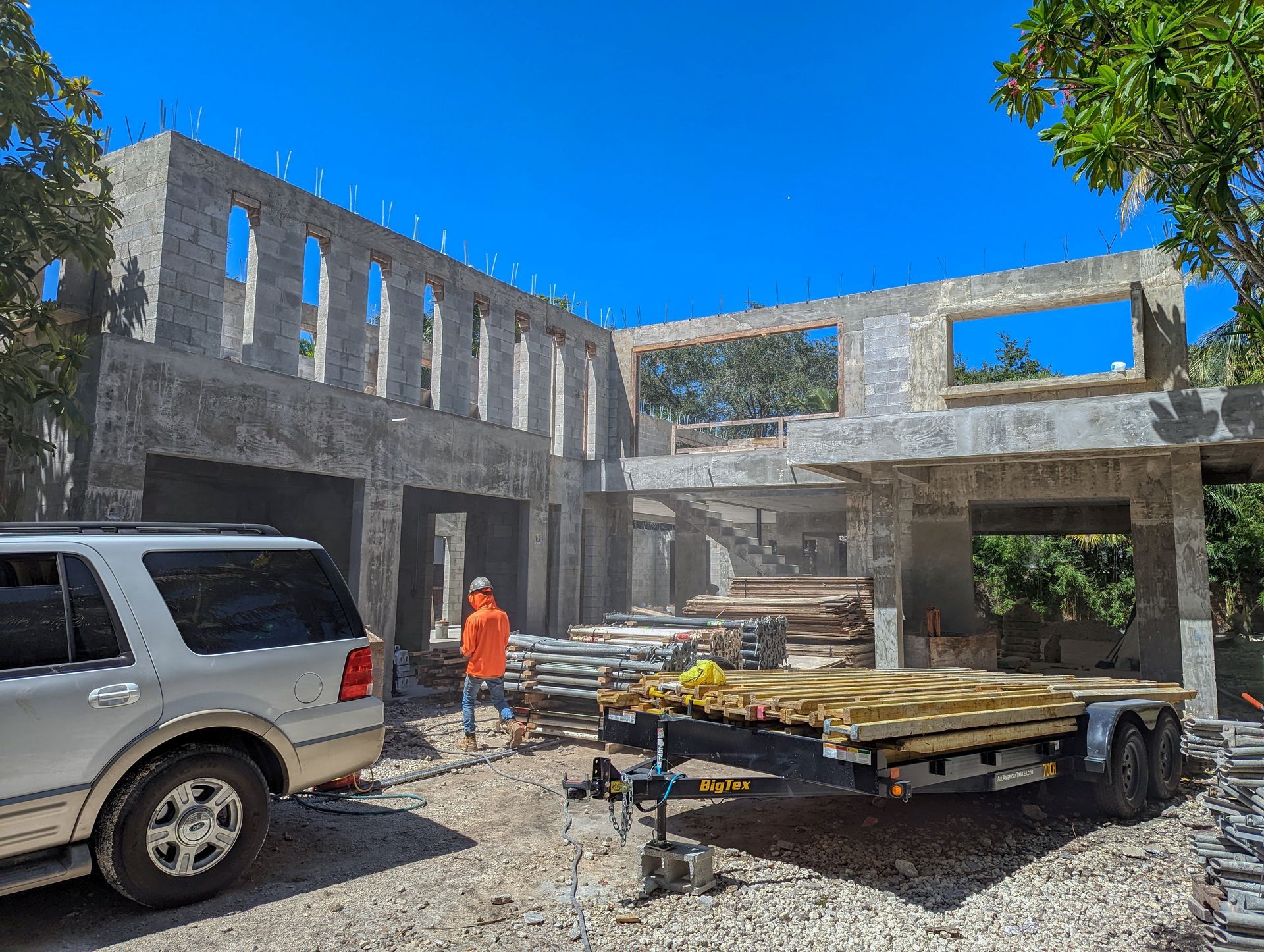 Construction site: partially built concrete house with an SUV, trailer, and worker.