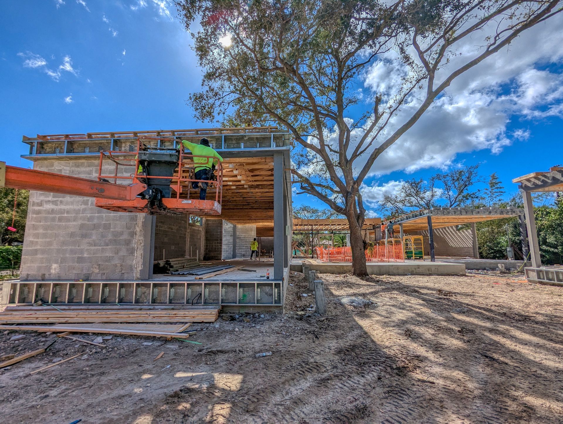 Construction site: Workers on lift building a concrete block structure with a large tree nearby.