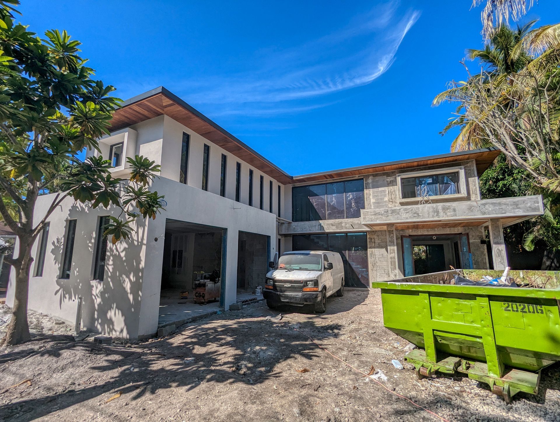 Two-story house under construction with a white van in the driveway and a green dumpster. Blue sky.