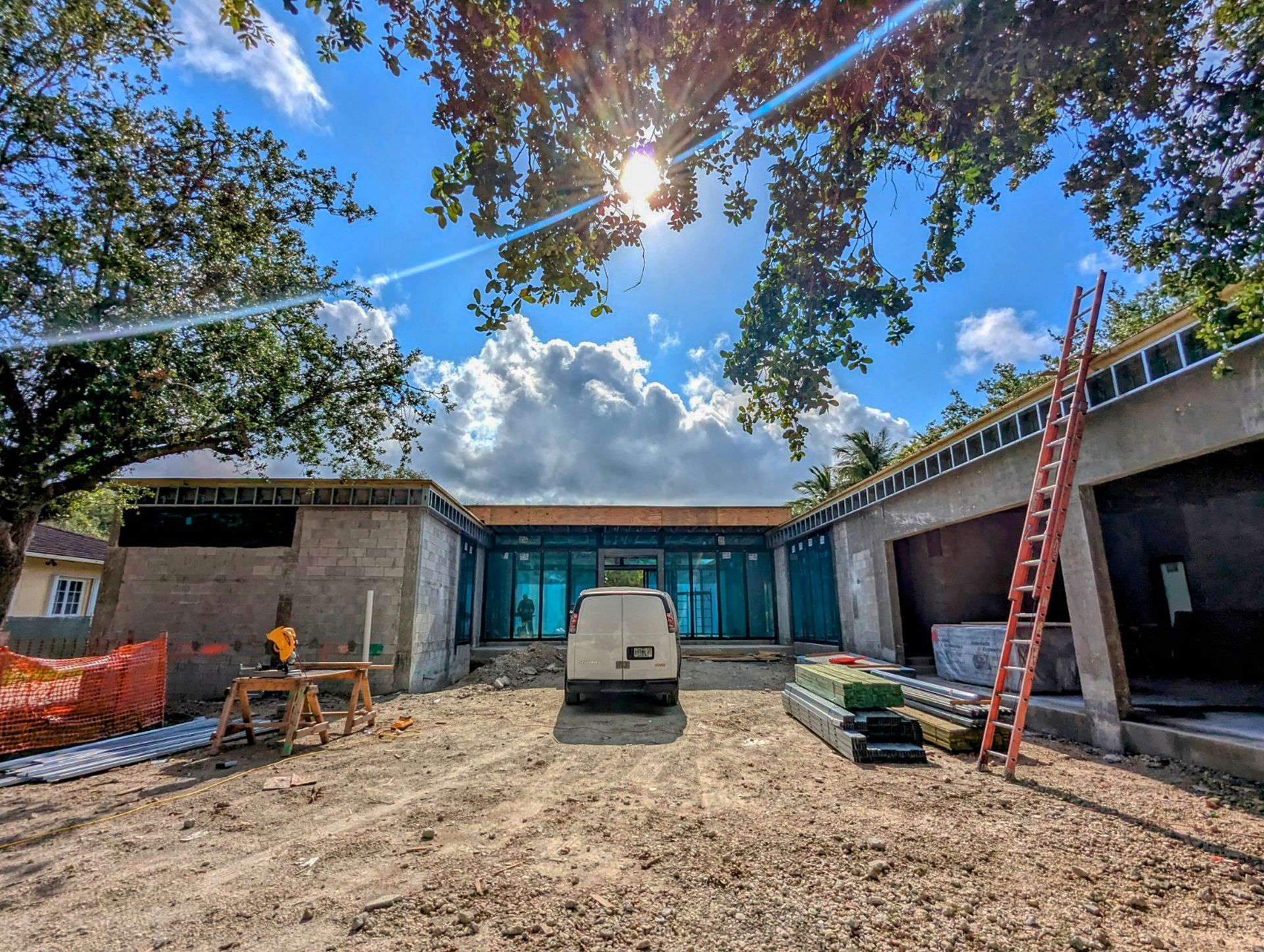 Construction site, partially-built building with a van parked in the center, bright sun, blue sky, trees.