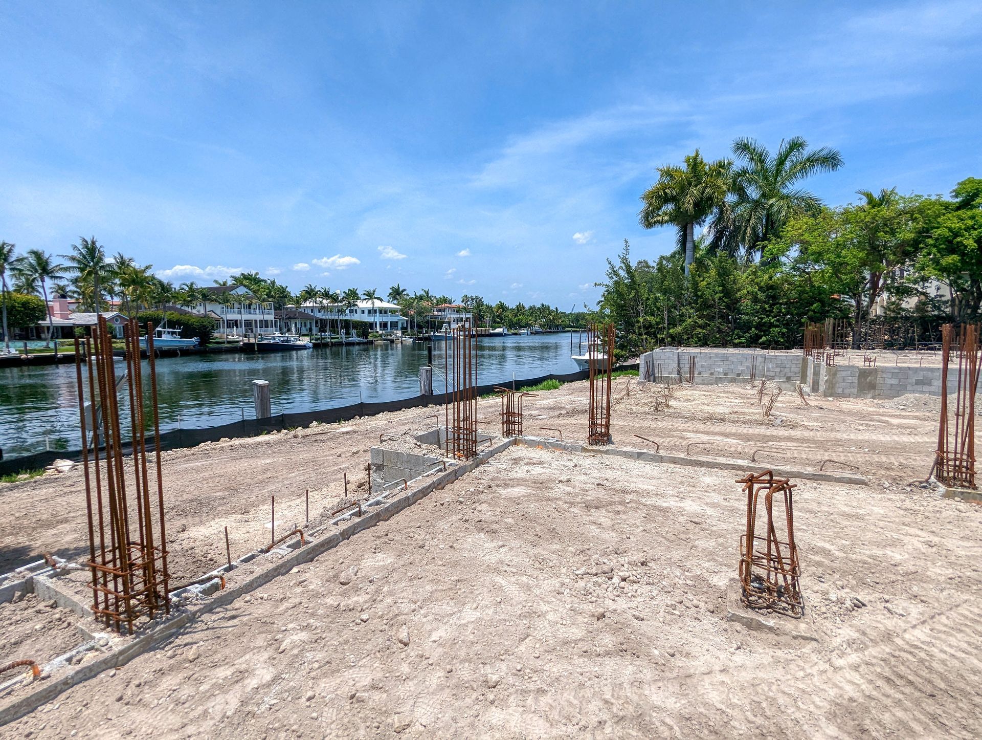 Construction site with concrete foundation and rebar, waterfront view with houses and trees.
