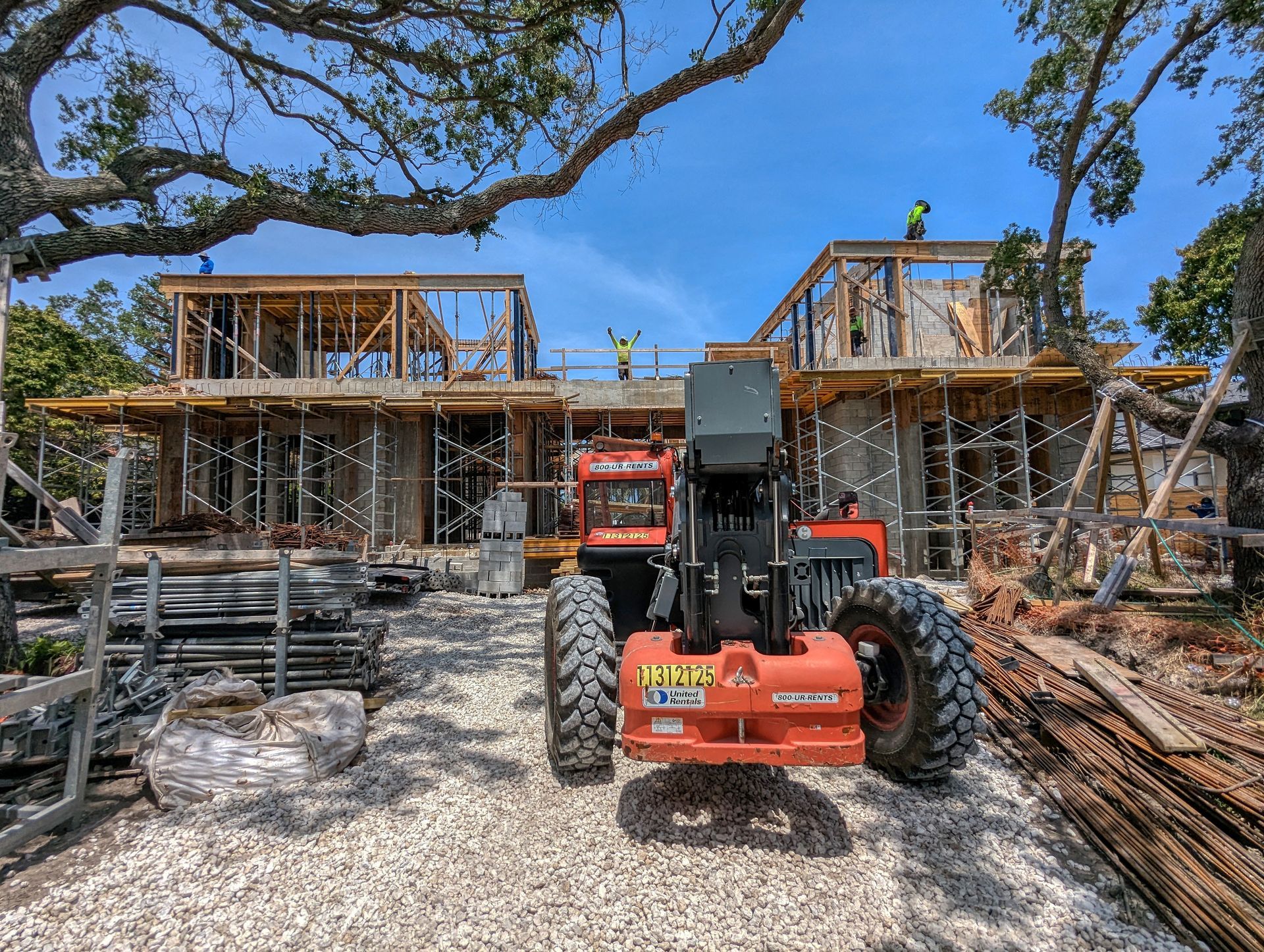 Construction site with an orange forklift in front of a partially built house, workers on roof, blue sky.